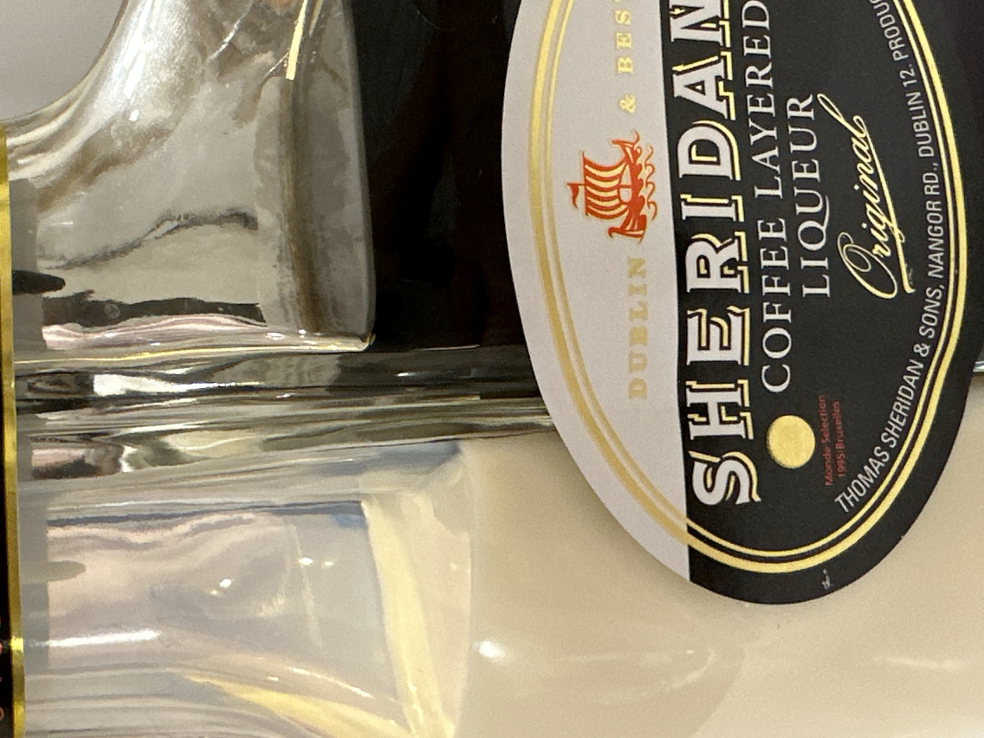 The image shows a close-up of a bottle of Sheridan's Coffee Layered Liqueur. The bottle is distinctive, with two separate compartments, one containing a white, creamy liqueur and the other a dark, coffee-colored liqueur. A black label with gold lettering is prominent, displaying the brand name "SHERIDAN'S", "COFFEE LAYERED LIQUEUR", and "Original". Below this, the label details "Monde Selection 1995 Bruxelles" and "THOMAS SHERIDAN & SONS, NANGOR RD., DUBLIN 12. PRODUC". The background is blurred, suggesting the bottle is the primary subject. The lighting is bright, with reflections visible on the glass bottle. The location is not discernible from the image.