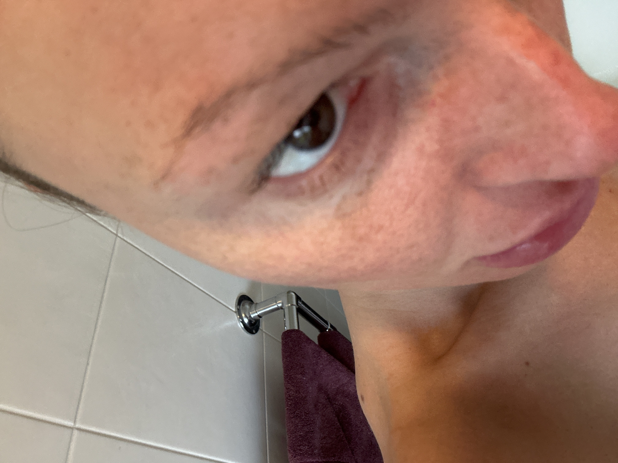 A close-up selfie of a person's face taken from a low angle. The person has light skin, brown hair, and brown eyes. The focus is primarily on the right side of the face, including the eye, nose, and mouth. The background shows a tiled wall, a towel rack, and a maroon-colored towel. There is no information in the image that identifies the location as being in Wealden, United Kingdom.