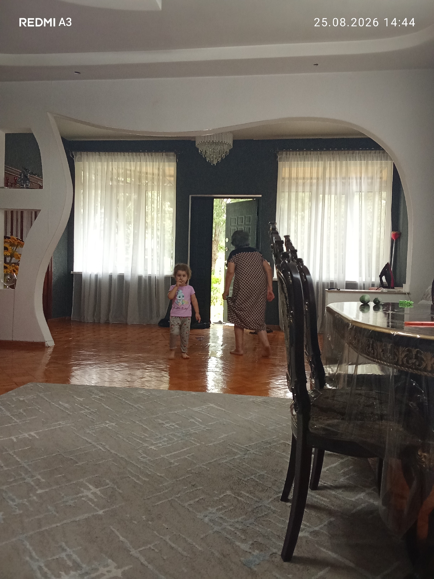 An interior scene shows a young child in a pink t-shirt and patterned pants walking on a polished wooden floor. Behind the child, a woman in a patterned dress is walking towards an open doorway. Large white curtains are visible on a wall behind them. In the foreground, a dark wooden chair with ornate carvings is positioned next to a table covered with a clear plastic cloth. The room has a high ceiling with an arched architectural feature. The timestamp "25.08.2026 14:44" and "REDMI A3" are overlaid on the image, indicating it was taken on August 25, 2026, at 2:44 PM. The location is identified as Pshap, Abkhazia, but this information is not visually verifiable from the image itself.