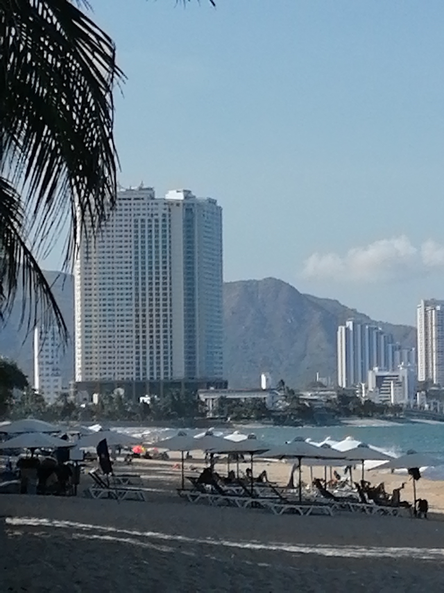 The image depicts a beach scene in Nha Trang Ward, Vietnam. A tall building stands in the background, with mountains behind it. Several other buildings are also visible in the distance. In the foreground, there is a beach with numerous lounge chairs and umbrellas. Some people are relaxing under the umbrellas. Part of a palm tree is visible on the left side of the image. The sky is light blue with a few white clouds.
