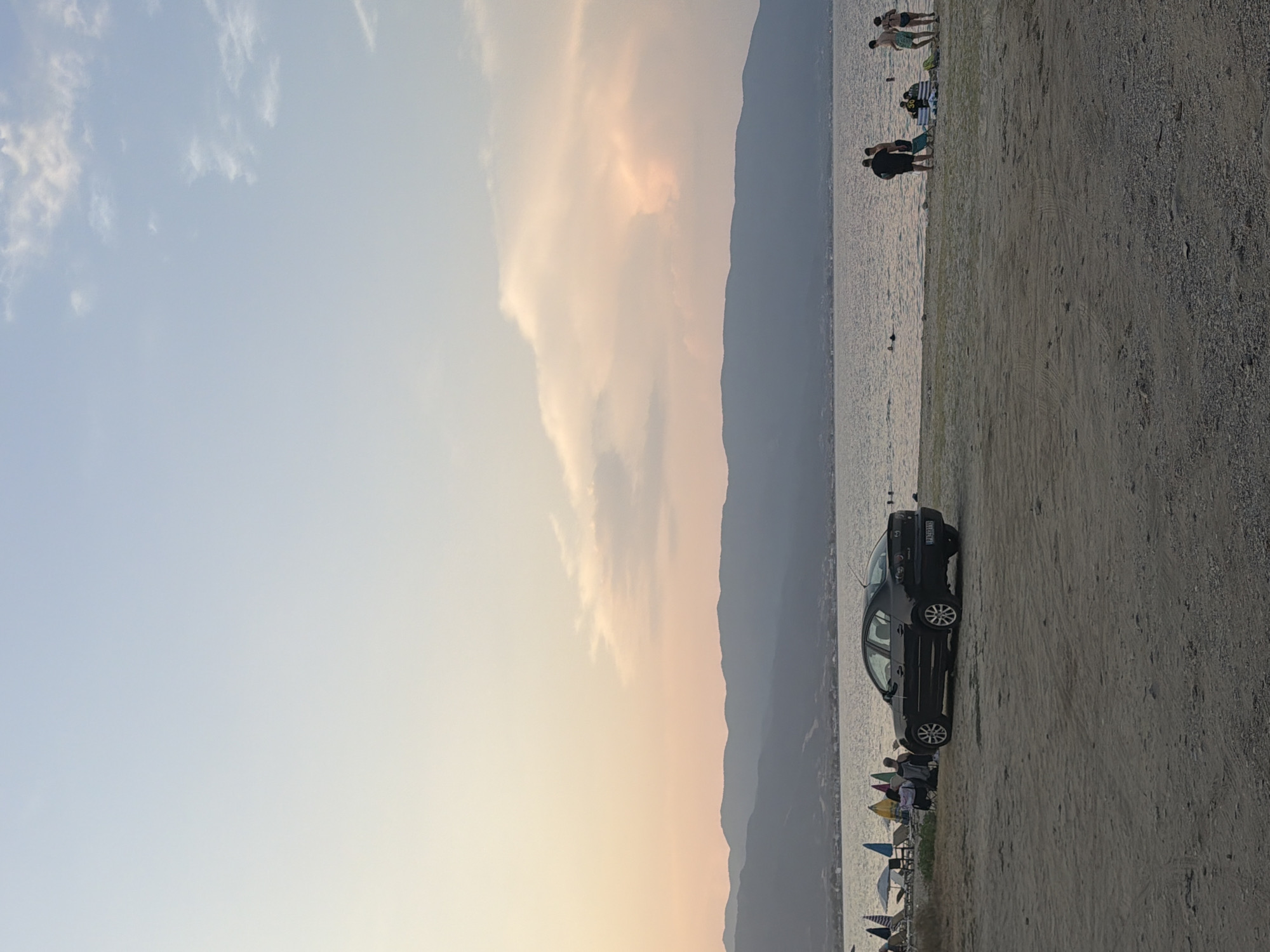 This image captures a serene sunset scene at a beach in Stavros, Greece. The sky transitions from a pale blue at the top to hues of pink and orange near the horizon, with wispy clouds illuminated by the fading sunlight. In the foreground, a sandy beach is sparsely populated. A dark-colored sedan is parked on the sand, facing towards the left. Further down the beach, several beach umbrellas and chairs are set up. A group of people are visible near the water's edge; some are standing and conversing, while others are in the shallow water. In the background, a calm body of water stretches out to meet a range of silhouetted mountains that form the coastline. The overall atmosphere is peaceful and recreational.