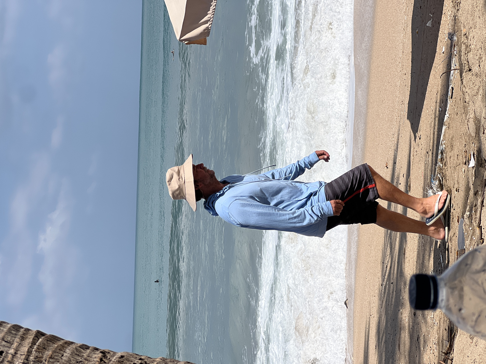A man wearing a blue hoodie, black and red shorts, a tan hat, and flip-flops walks along the beach in Kuta, Indonesia. The man is walking from left to right and his body is partially obscured by a palm tree trunk on the left side of the frame. The ocean is to his right, with waves breaking on the shore. In the distance, there are two people in the water, possibly swimming or surfing. A beige umbrella is visible on the far right of the image. The sand on the beach is light brown and there are some scattered pieces of debris. A partially visible plastic water bottle is in the foreground on the left. The sky is a pale blue with some scattered clouds.