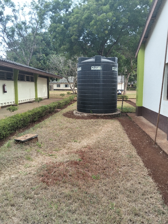 A large black water tank sits on a concrete pad between two buildings with light-colored walls and green trim. The surrounding grassy area is mostly dry, with patches of bare soil. A low hedge runs along the side of one building. A covered concrete access point is visible in the foreground. Trees provide a backdrop, and another building can be seen in the distance. The sky appears overcast.