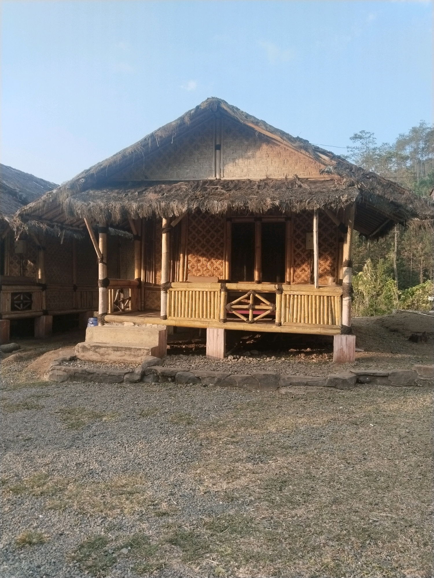 A traditional Indonesian bamboo hut with a thatched roof is shown in the foreground, bathed in natural light. The hut is elevated on stilts and features intricate woven bamboo walls and a wooden railing. A small set of concrete steps leads up to the entrance, which has glass doors. The ground around the hut is covered with gravel and patches of grass. In the background, a dense forest with tall trees is visible under a clear blue sky. The scene appears to be located in Guci, Indonesia, a popular tourist destination known for its natural hot springs and scenic landscapes. There are no people present in the image.