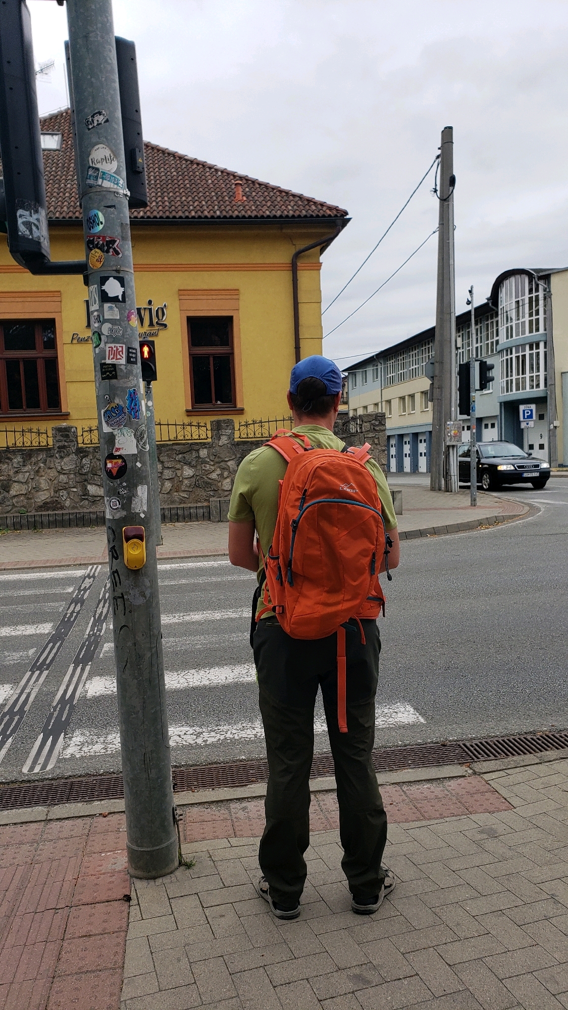 The image shows a person with an orange backpack standing on a sidewalk next to a pedestrian crossing in the city of Prešov, Slovakia. The person is facing away from the camera and is wearing a green shirt, dark pants, and sandals. A traffic light pole with stickers and a pedestrian button is to the left of the person. In the background, there is a yellow building with a red tiled roof, a stone wall, and modern buildings across the street. A black Audi car is visible in the distance. The sky is overcast. The location is identified as Prešov, Slovakia based on the street sign "ZÓNA P" visible in the background, which is a common parking zone sign in Slovakia.