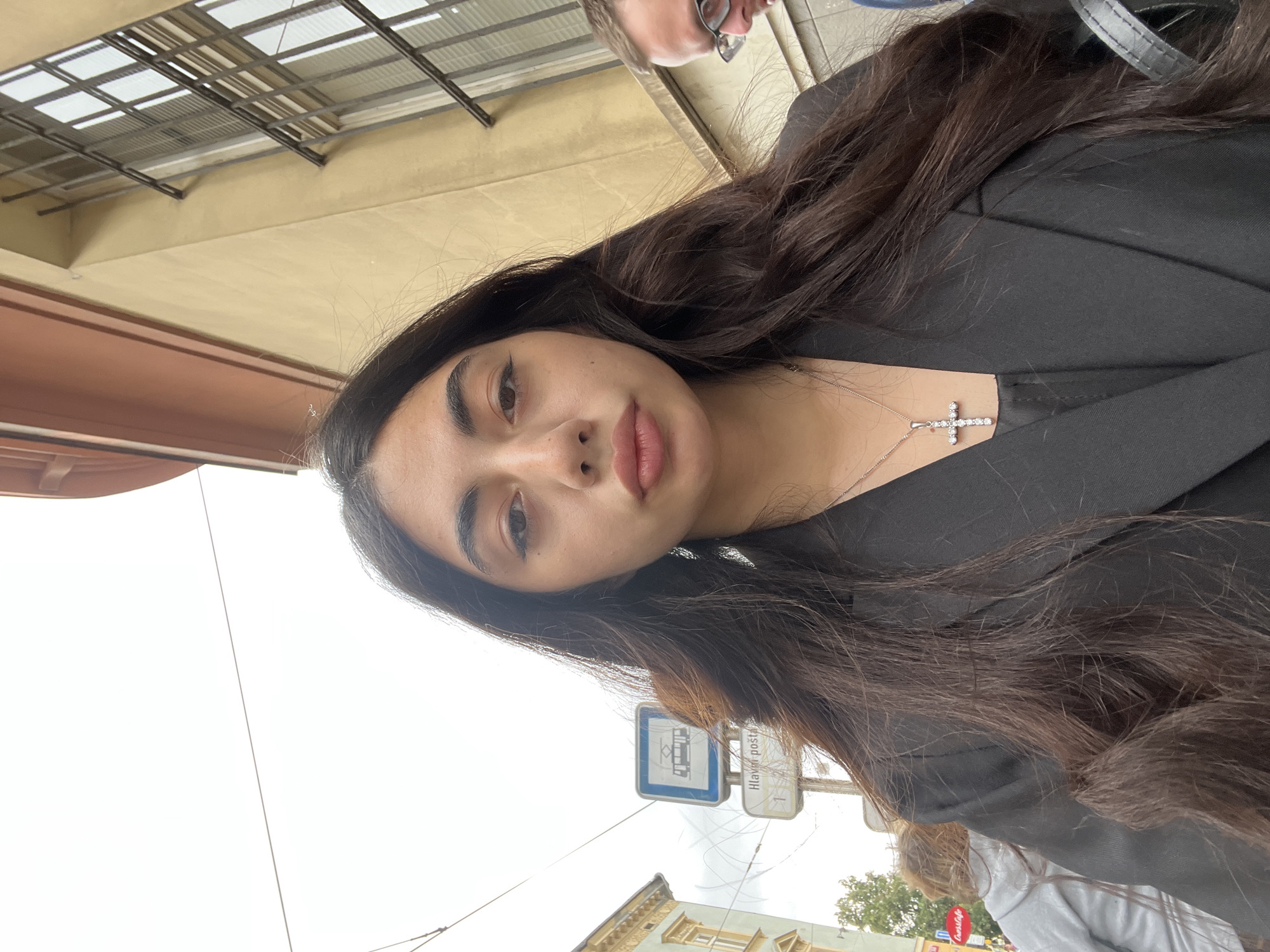 A medium-shot selfie, taken from a low angle, shows a young woman with long, dark, wavy hair and fair skin. She is wearing a black blazer over a dark top and a silver cross pendant necklace. Her eyes are looking directly at the camera. In the background, a street scene is visible, including a tram stop sign with the text "Hlavní pošta" and the number "1", indicating it is the main post office stop. The architecture in the background suggests a European city, possibly Pilsen, Czechia, with its characteristic building styles. A man with glasses is partially visible to the right of the woman, and other people and a red "Coop" store sign are visible in the distance on the left. The sky is overcast.