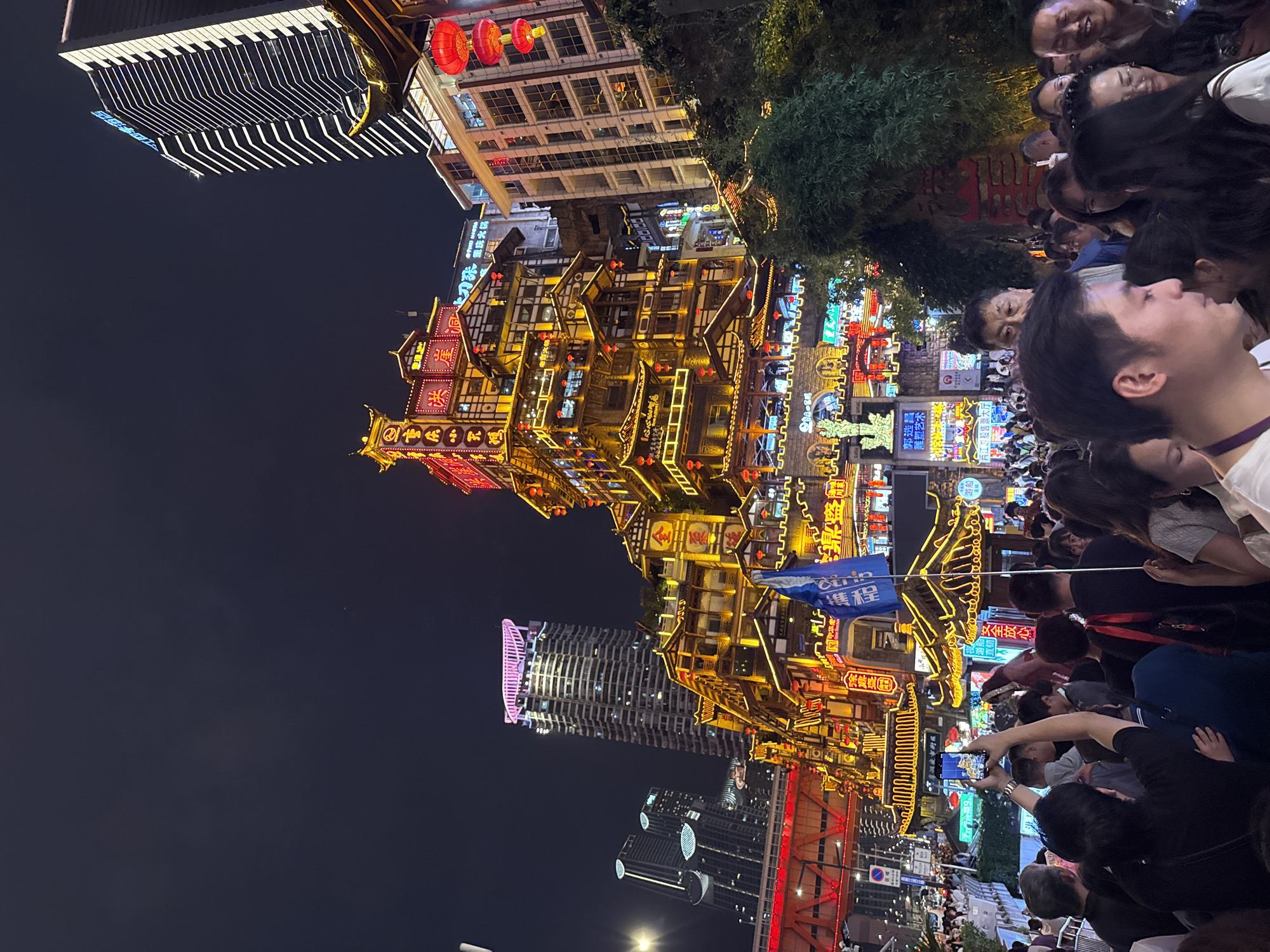 The photograph captures a bustling night scene at the Hongyadong tourist area in the Yuzhong District of Chongqing, China. The focal point is a towering, multi-tiered architectural structure, illuminated with an array of warm-toned lights and red lanterns, giving it a traditional Chinese appearance. The architecture is reflected in a river, which is partially obscured by modern buildings.
A dense crowd of people gathers near the foreground, with many holding up smartphones, presumably to capture images of the illuminated architecture. The atmosphere suggests a popular tourist destination. There are signs in Chinese advertising services, vendors, and tourist information. The sky is dark, indicating it is nighttime, which accentuates the vibrant lighting of the buildings and structures.