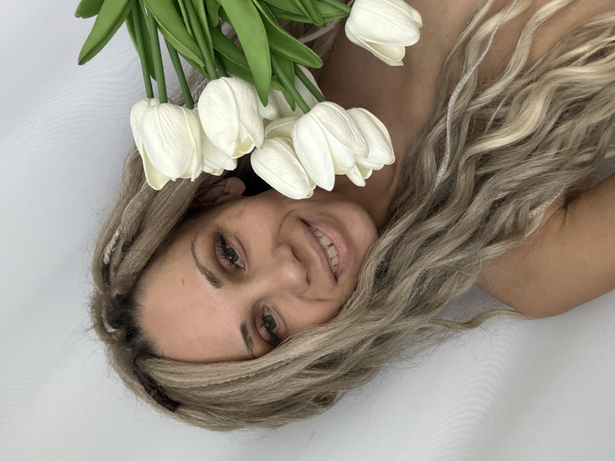 A woman with light blonde hair is lying on a white surface, holding a bouquet of white tulips near her face. Her head is tilted to the side, and she is smiling. The background is a plain white. The location, Rusanovo, Russia, is not discernable from the visual information provided.