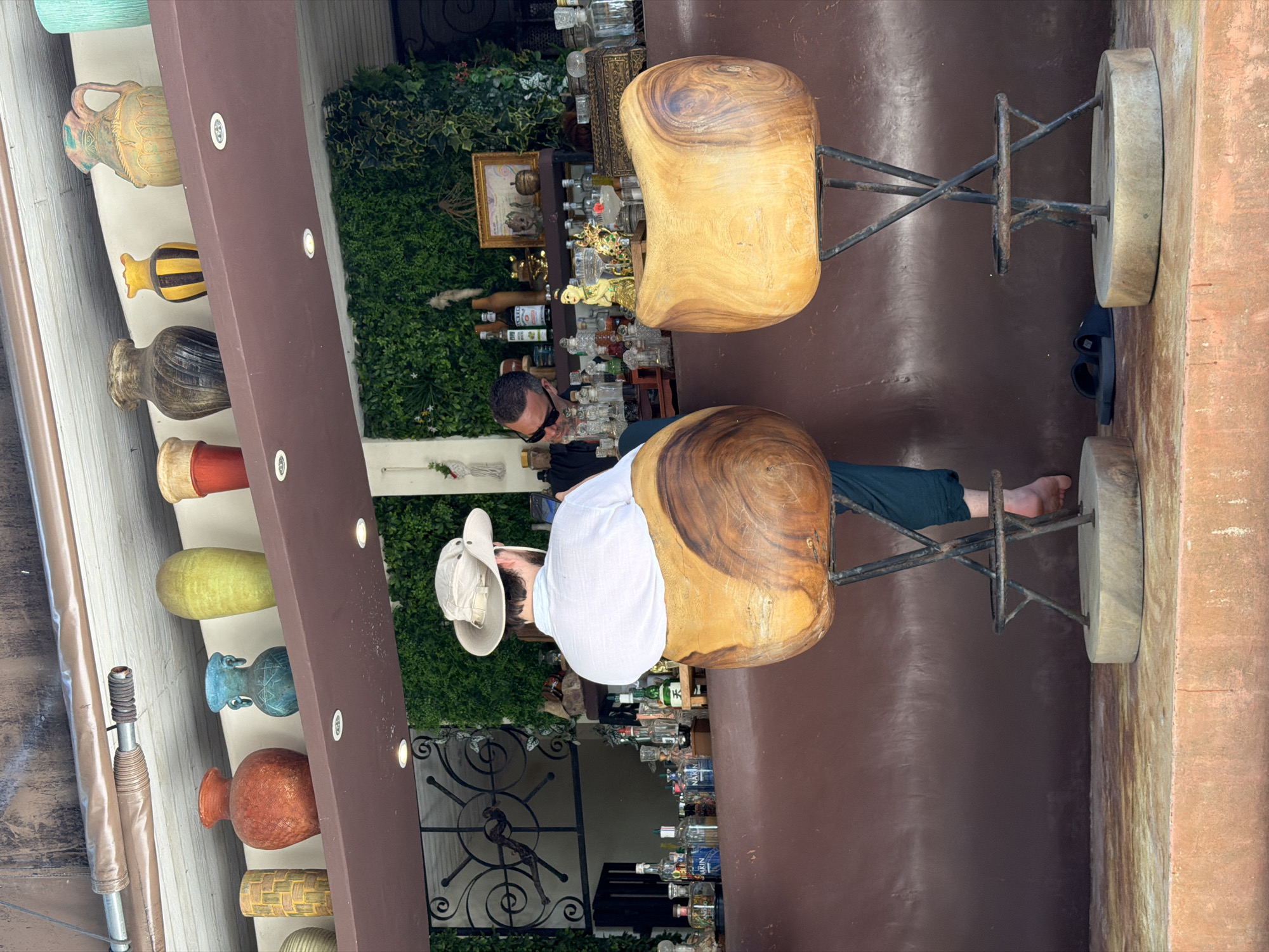 The image depicts a man wearing a white hat and white shirt sitting on a stool in what appears to be an outdoor establishment, possibly a bar or restaurant, in Baan Thong Krut, Thailand. The man is facing away from the camera, his body angled to the right. He is seated on a large, carved wooden stool with a metal base. In the background, shelves are filled with various bottles, suggesting a bar setting. There is also a lush green wall of foliage, a common decorative element in tropical locations. The overall scene suggests a relaxed atmosphere, potentially during the daytime given the brightness. The man's posture and attire are casual. The image is taken from a slightly elevated perspective, looking down and to the side. The overall color palette is warm, with earthy tones from the wood and building materials contrasted by the vibrant green of the plants and the varied colors of the bottles.