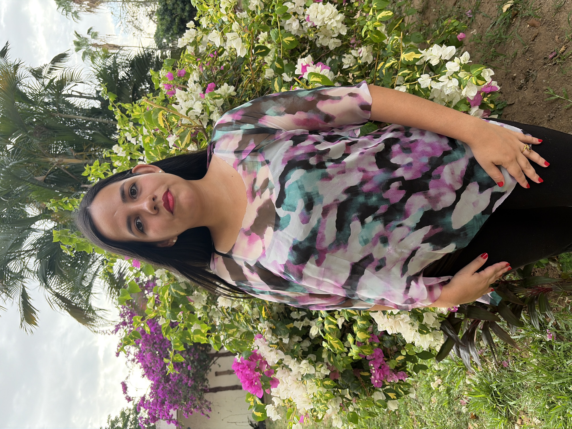 A woman stands in a garden setting. She has dark hair, red lipstick, and red nail polish. She wears a semi-sheer, patterned top with shades of white, pink, teal, and brown, along with black pants. She is wearing a ring on her right hand. The background includes a lush bougainvillea bush with vibrant pink and white flowers, along with other greenery and palm trees. The sky above is partly cloudy. The location is Ciudad Guayana, Venezuela.