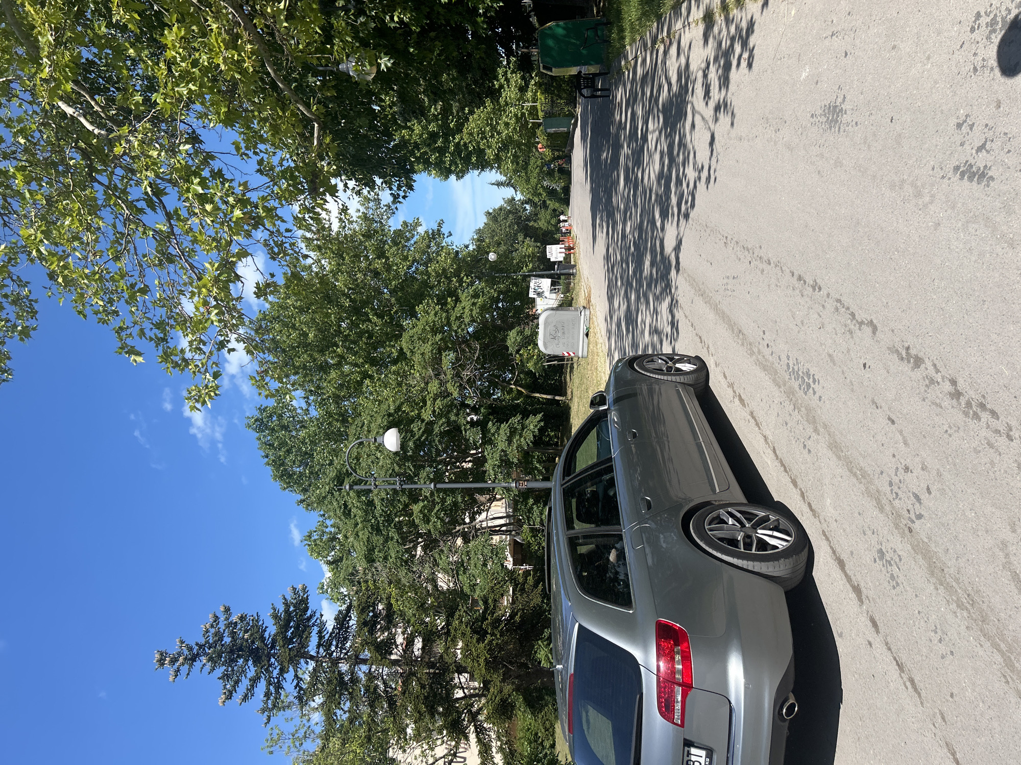 In Varna, Bulgaria, on a sunny day, a grey Audi A6 Avant is parked on the right side of a paved road. The car is positioned on the left lane, facing away from the viewer. The road is bordered by lush green trees on both sides, casting dappled shadows on the asphalt. In the distance, the road curves gently to the right, leading towards more trees and a clear blue sky with scattered white clouds. Several streetlights are visible along the roadside, and a few people can be seen walking in the distance. A dark green trash bin and a black bench are also present on the right side of the road. The overall scene suggests a peaceful, park-like setting within the city.