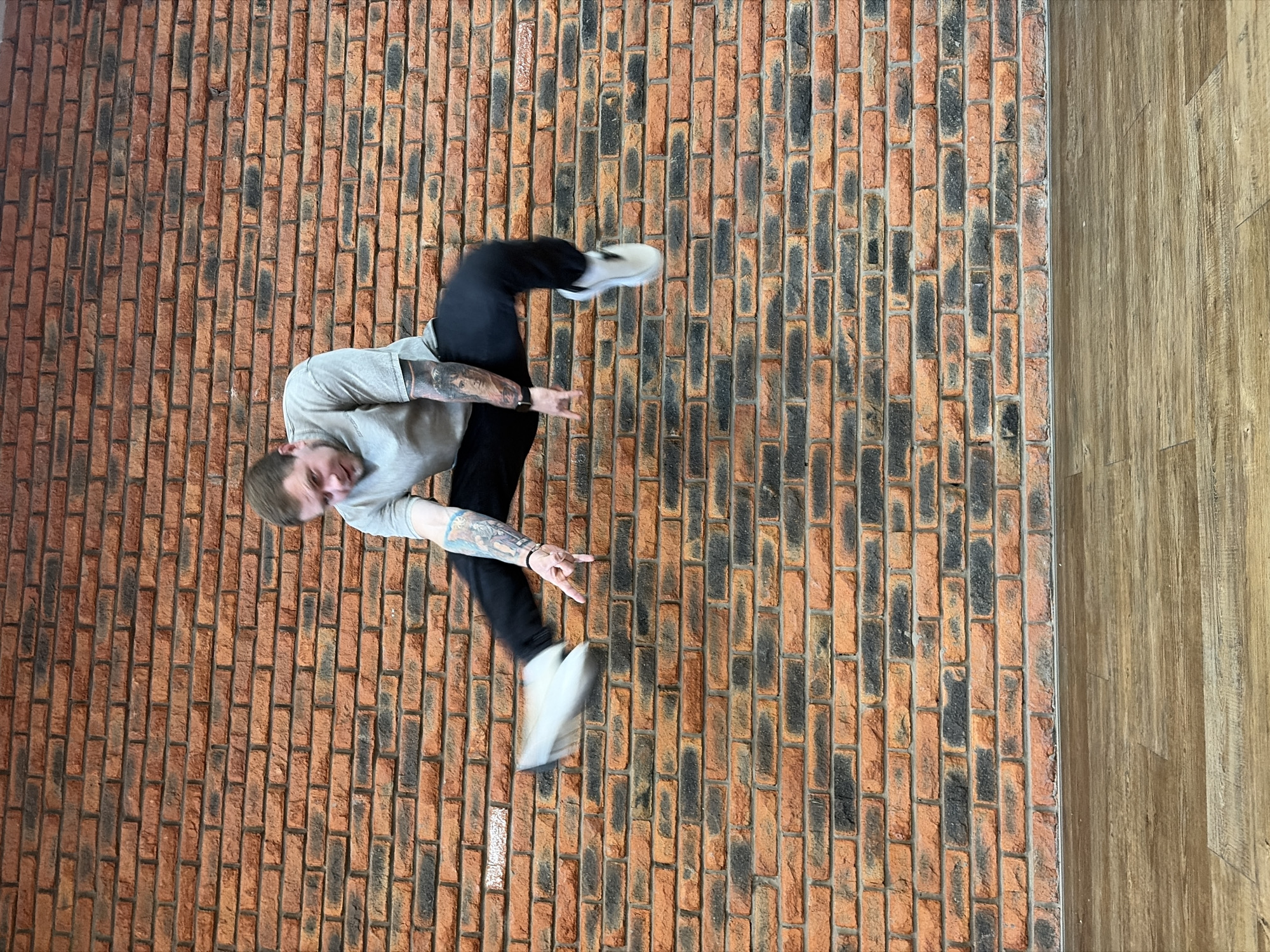 The image displays a dynamic indoor scene in Chaikovsky, Russia, featuring a man in mid-air against a textured background. The primary subject is a man, dressed in a light grey t-shirt, dark pants, and white sneakers, with visible tattoos on both forearms. He is captured horizontally across the frame, appearing to be jumping or performing an acrobatic move, with his body bent and legs lifted. His right hand is extended in a "rock on" gesture. His face is visible, showing a focused or neutral expression, and his hair is light brown. The blurring around his feet indicates motion.
The setting consists mainly of a prominent brick wall, characterized by a pattern of orange-red and dark grey bricks. To the far right of the image, a section of light brown wooden flooring is visible, running vertically parallel to the brick wall. The lighting is bright and even, suggesting an indoor environment, possibly a studio or a modern room. There are no specific cues for the time of day or weather, apart from the indoor setting. No visible text is present in the image. The overall impression conveys energy and movement within a contemporary, industrial-chic interior.