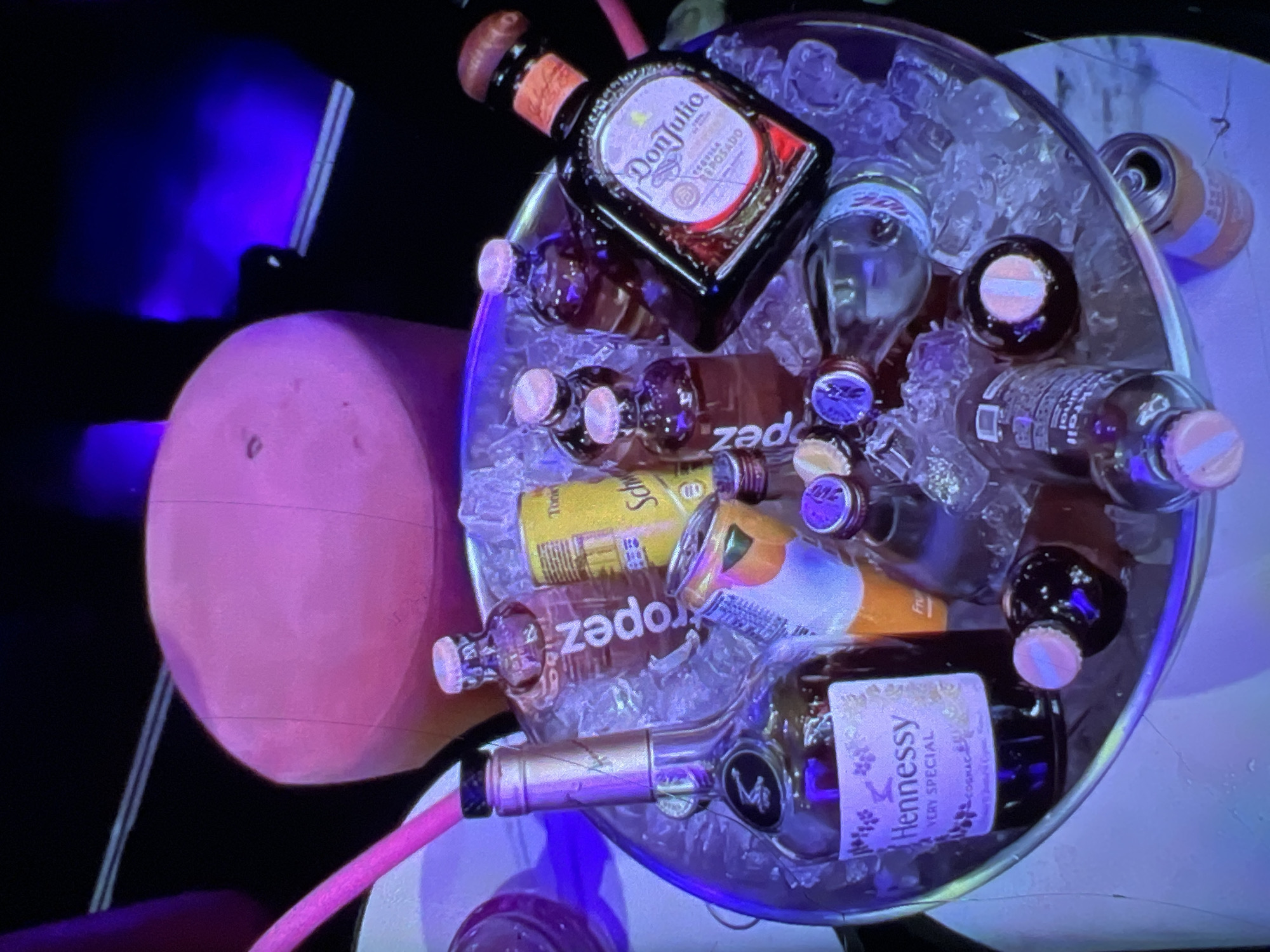 In a dimly lit room, possibly a VIP section of a nightclub in Lagos, Nigeria, an ice-filled bucket contains an assortment of alcoholic beverages. Prominently displayed is a bottle of Hennessy Very Special Cognac and a bottle of Don Julio Tequila. Several smaller bottles, some with pink caps, are also present, along with a can of Schweppes Tonic Water and another can that appears to be orange-flavored. A pink straw extends from the bucket. The background features a large, plush, pink circular stool and a wall illuminated with blue lights, creating a vibrant and celebratory atmosphere. There are no visible people in the image.