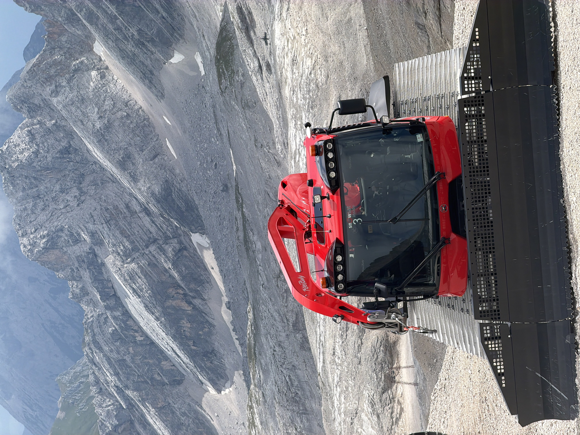 A bright red snow groomer is positioned on a rocky, mountainous terrain. The snow groomer has a large blade at the front and a robotic arm extending from the top. The cabin of the snow groomer is visible, with several lights and a windshield. The background consists of rugged, grey mountains with patches of snow and scree. In the distance, a small, dark object resembling an airplane or drone is visible in the sky. The overall scene suggests a high-altitude environment, likely a ski resort area in the Alps, possibly near Garmisch-Partenkirchen, Germany, known for its mountainous landscape. The ground beneath the snow groomer is covered in gravel and small rocks.