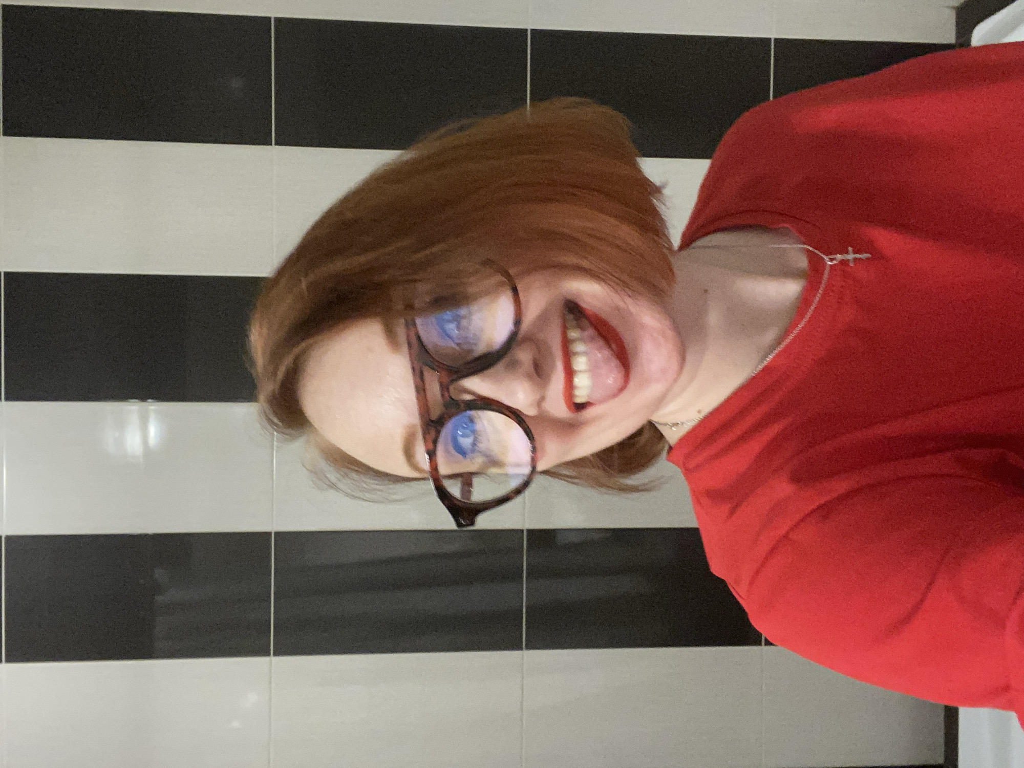 A person with reddish-brown hair, wearing glasses and a red shirt, sticks their tongue out towards the camera. They also have a silver necklace. The background features black and white striped tiles. The location of the image is unclear and could not be forensically determined as Yekaterinburg, Russia.