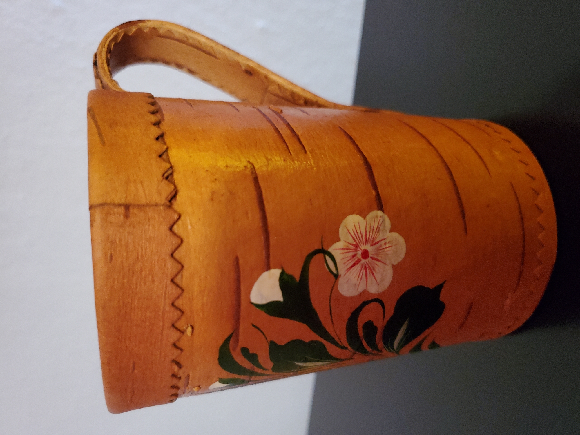 This is a medium close-up, eye-level, indoor shot of a wooden mug with a handle. The mug is a light brown color with a natural wood grain texture. It has a decorative zigzag pattern stitched around the top rim and a similar pattern stitched around the bottom rim. There are horizontal lines carved into the side of the mug, giving it the appearance of bark. A floral design is painted on the side of the mug, featuring green leaves and two white and pink flowers. The mug is placed on a dark, reflective surface. The background is a plain, light-colored wall. The image does not provide any information or context related to Konstantinovka, Moldova.