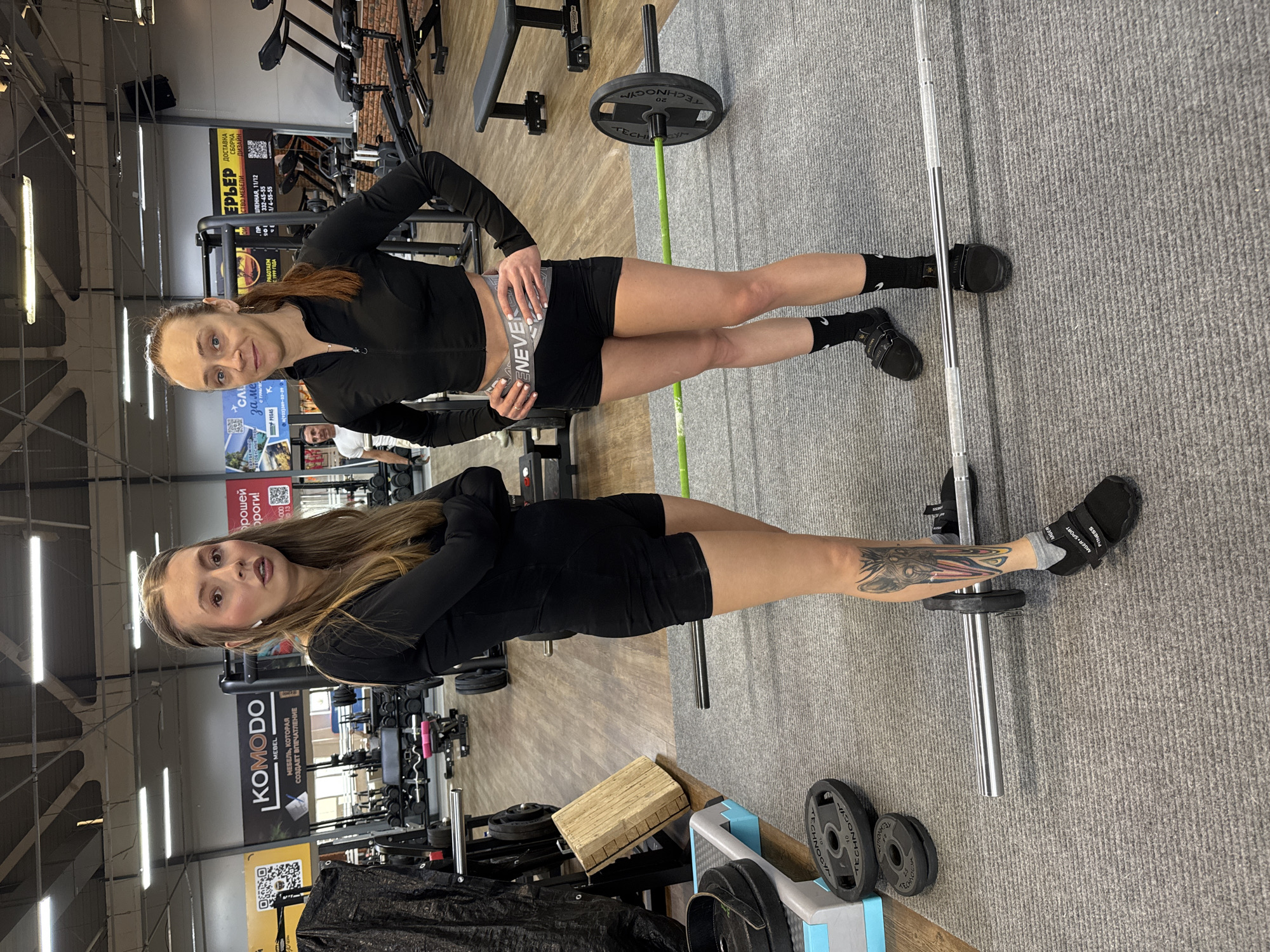 Here is a detailed description of the image:
The image depicts two young women in what appears to be a fitness center or gym. They are standing side-by-side, facing the camera.
The woman on the right has auburn hair pulled back in a ponytail, wearing a black zip-up athletic jacket and black shorts. She is holding a water bottle. The woman on the left has long, light brown hair and wears a similar outfit. She has a tattoo on her left leg.
Both women are standing near gym equipment. Visible in the background and foreground are weight plates, barbells, and various workout machines. The floor is covered with gray carpeting, and the lighting suggests a commercial or public space. The gym features include “KOMODO” branding, suggesting a commercial gym chain. Signage and text in Russian indicate a location potentially in Russia or a Russian-speaking region.