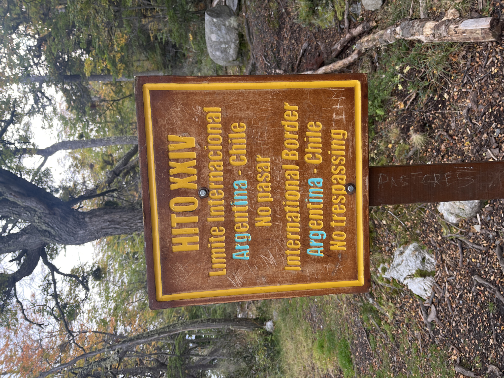 The image shows a weathered, brown and yellow sign marking the international border between Argentina and Chile. The sign, titled "HITO XXIV," displays text in both Spanish and English, indicating "Limite Internacional Argentina - Chile" and "International Border Argentina - Chile," along with "No pasar" and "No trespassing." The sign is mounted on a brown wooden post, and the word "PASTORES" is faintly scratched into the post below. The sign is situated in a natural environment with trees and ground covered in leaf litter and vegetation. The surrounding environment suggests a forested area, possibly a park or hiking trail. The image is taken at ground level, and the perspective is slightly tilted. The location is Lapataia, Argentina.