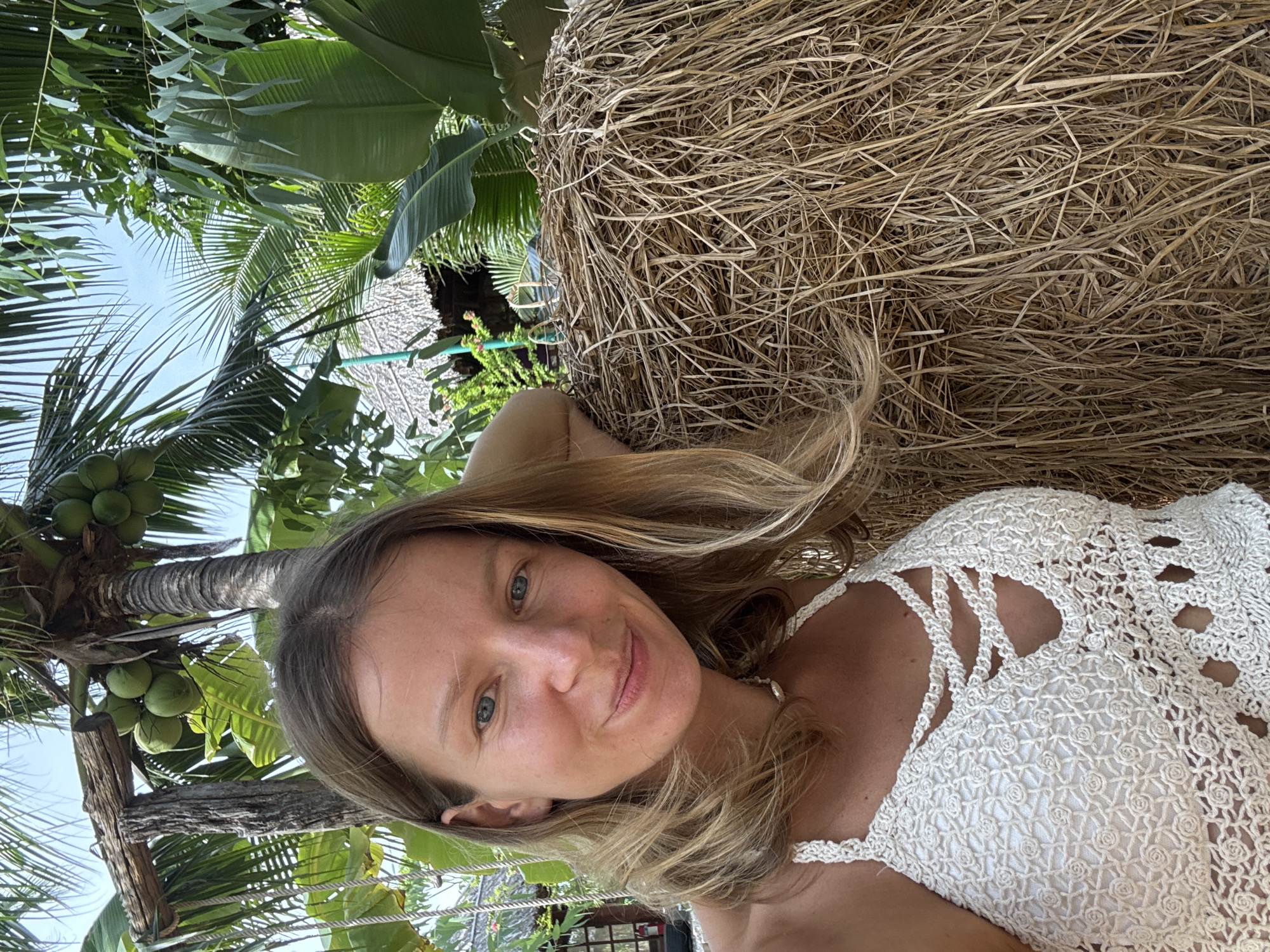 A blonde woman with blue eyes smiles at the camera, leaning on a large bale of hay. She is wearing a white crocheted top. In the background, palm trees with coconuts are visible, along with a thatched roof structure and dense tropical foliage. The setting appears to be a resort or natural landscape in Mũi Né, Vietnam.