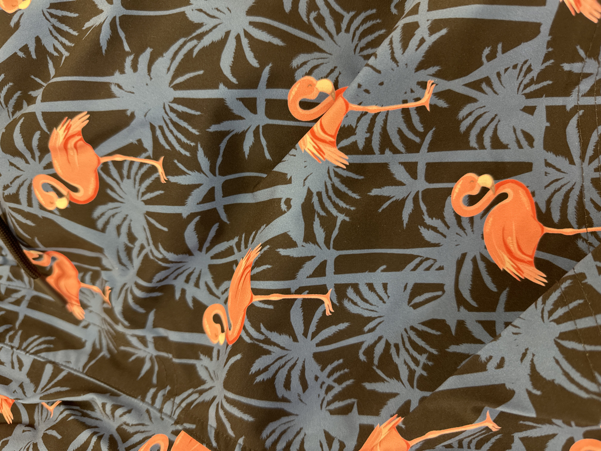 The image is a close-up of a fabric with a tropical pattern. The pattern consists of stylized orange flamingos and silhouettes of palm trees against a dark background with blue palm fronds. The fabric appears to be part of a garment, possibly swimwear, as suggested by the drawstring visible at the top left. There are no people or specific locations within Steenwijk, Netherlands, identifiable in this image.