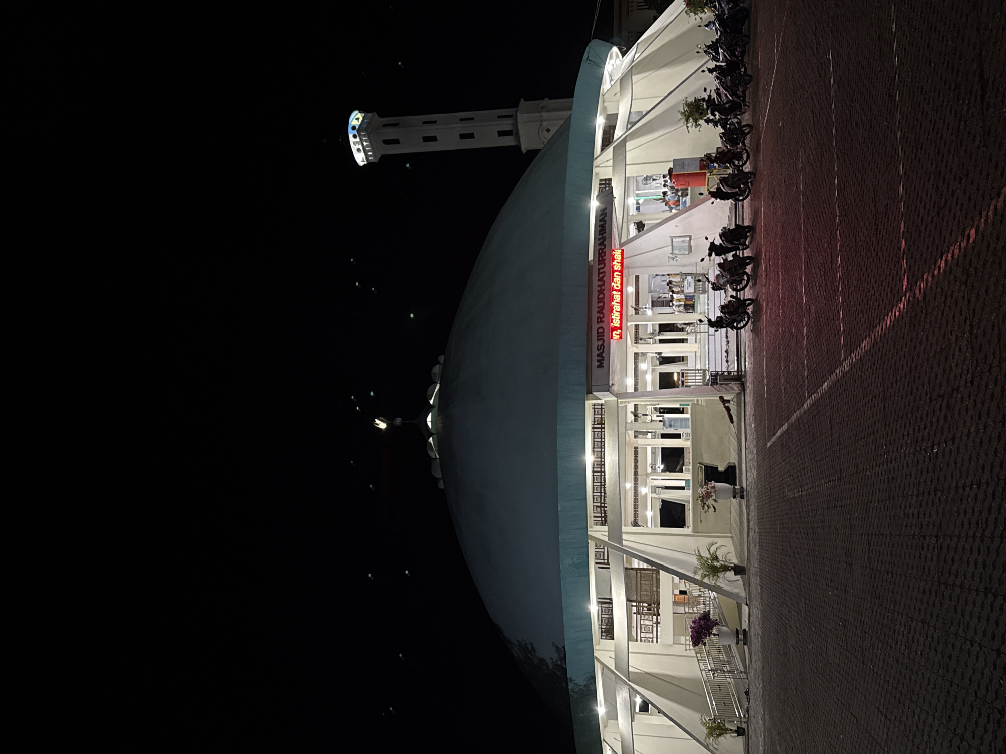 Here's a description of the image:
The image is taken at night of the Masjid Raudhaturrahmah, a mosque in Pasar Paloh, Indonesia. The mosque has a large dome and a tall minaret. A brightly lit LED sign with red lettering is visible on the mosque's facade. Motorbikes are parked in a row along the mosque's edge on a brick paved surface. The sky is black.