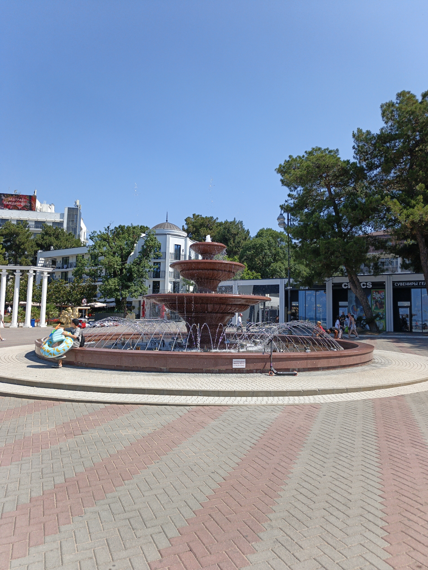 A tiered fountain with red stone bowls is the centerpiece of this outdoor scene, surrounded by a paved area with a distinctive red and grey herringbone pattern. Water sprays from the fountain, catching the bright sunlight.
In the foreground, a young person wearing a black cap sits on the edge of the fountain, holding a blue and yellow inflatable ring. Behind them, several people are gathered near the fountain and walking on the paved area.
Further back, a row of shops with glass storefronts lines a street. Signs visible on the shops include "CROCS" and "СУВЕНИРЫ ГЕЛ". Green trees with full canopies are abundant, some casting shadows on the buildings and pavement. In the background, white multi-story buildings with balconies suggest a resort or urban area. A white, colonnaded structure stands to the left of the fountain.
The sky is a clear, bright blue, indicating a sunny day, likely mid-morning or early afternoon given the strong shadows. The overall atmosphere is one of a pleasant, warm day in a public outdoor space, possibly a promenade or town square in a location like Pshada, Russia. A small black electric scooter is parked near the base of the fountain. A sign with Russian text is attached to the fountain's edge. The text on the sign reads "ЗАПРЕЩЕНО МЫТЬ НОГИ НА МОНТАРЕ!" which translates to "It is forbidden to wash your feet on the counter!". Another sign on a building displays "ЗАВТРАКИ КАЖДЫЙ ДЕНЬ" meaning "Breakfast every day".