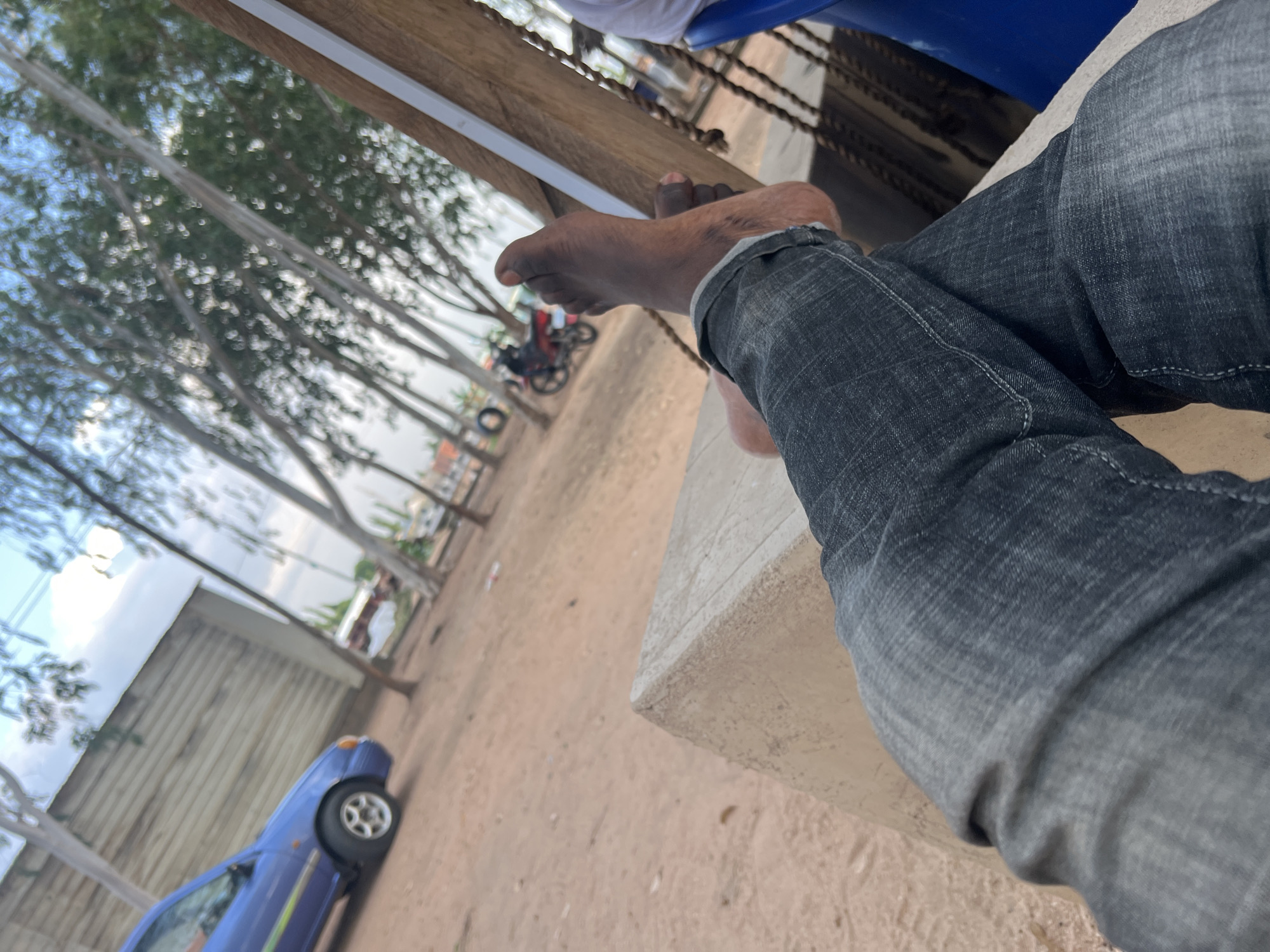 The image depicts a relaxed scene in an outdoor, sandy area, possibly a rest stop or a casual gathering spot in Mayo Dasa, Nigeria. In the foreground, a person's legs, clad in dark denim jeans, are crossed. The person's bare feet are visible, with the left foot resting on top of the right. One foot is dark-skinned. The person appears to be seated on a concrete structure.
In the mid-ground, a blue car is parked on the left, facing away from the viewer. Further back, a red motorcycle is parked, with a rider on it. The environment is characterized by a dirt ground with scattered debris. Tall, thin trees with sparse foliage line the area, creating a natural canopy overhead. A wooden structure with a white stripe runs vertically on the right side of the frame, suggesting it's part of a shelter or seating arrangement. In the background, there are buildings and more trees, indicating a semi-rural or a less developed urban setting. The sky above is partly cloudy. The overall impression is one of a casual, possibly humid, outdoor environment.