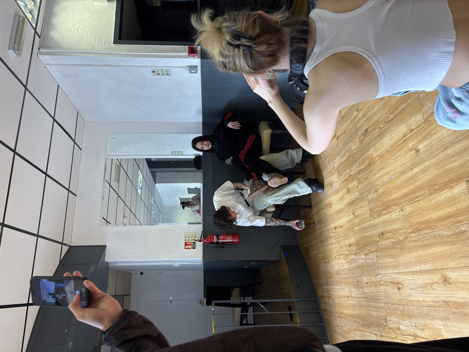 The image captures an indoor scene within a commercial establishment, possibly a former cafe or public house, located in London, United Kingdom. Four individuals are present, engaged in what appears to be a photography or video session.
In the immediate foreground, a person with blonde hair in a bun, wearing a white tank top and grey sweatpants, is crouched with their back to the viewer, holding a digital camera. To the left, a partial view shows another person's arm holding a black smartphone vertically, aimed towards the mid-ground.
The mid-ground features two individuals seated on light-colored chairs. The male on the left, displaying tattooed arms, wears a white t-shirt, light grey textured trousers, and dark shoes. He holds a glass containing an amber liquid in his left hand, resting it on the floor. A light-colored item is positioned on his right thigh. To his right, a female with long dark hair, wearing a black tracksuit with red side stripes, is seated with her left arm resting on the chair's back. Both seated individuals appear to be posing.
The background consists of white textured walls above a dark grey lower section, with a large rectangular mirror reflecting a person in a light shirt and dark cap further back in the room. A red fire extinguisher and a red fire alarm call point are visible on the wall. The ceiling features white acoustic tiles with a black grid pattern and integrated lighting. The floor is light brown laminate wood-style. A dark-framed opening or counter area is visible behind the seated subjects.