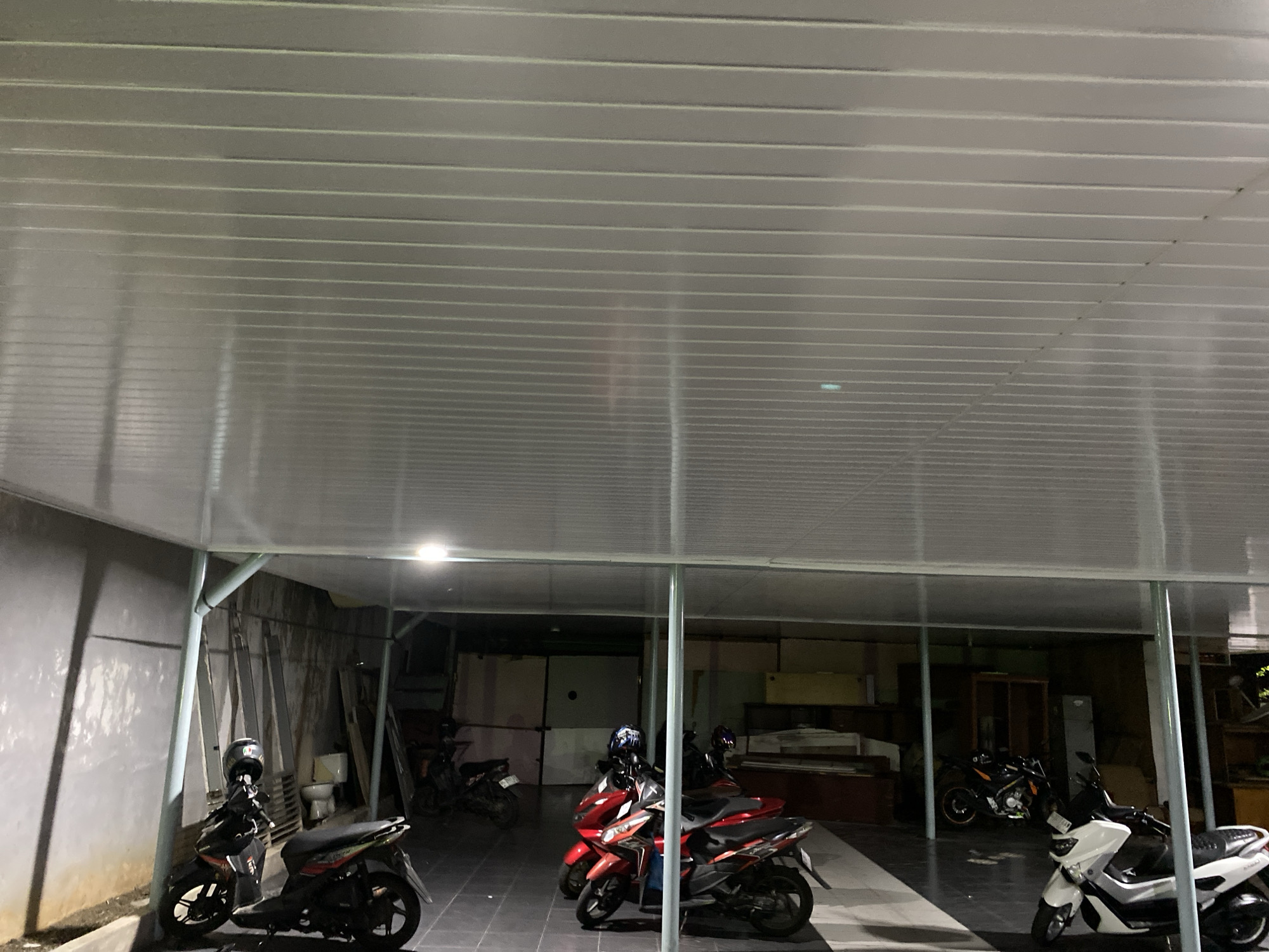 A covered parking area at night features several parked motorcycles. The ceiling is made of translucent, corrugated material with visible support beams. On the left, a black and red scooter is parked with a helmet on its handlebars. Further back, two red scooters are parked side-by-side. To the right, a white scooter is prominent, and behind it, a black and orange motorcycle is visible. The ground appears to be paved. The background shows a storage area with various items, including shelving, cabinets, and what looks like a toilet. A concrete wall with peeling paint is on the far left. The lighting in the scene is artificial, with overhead lights illuminating the parking area.