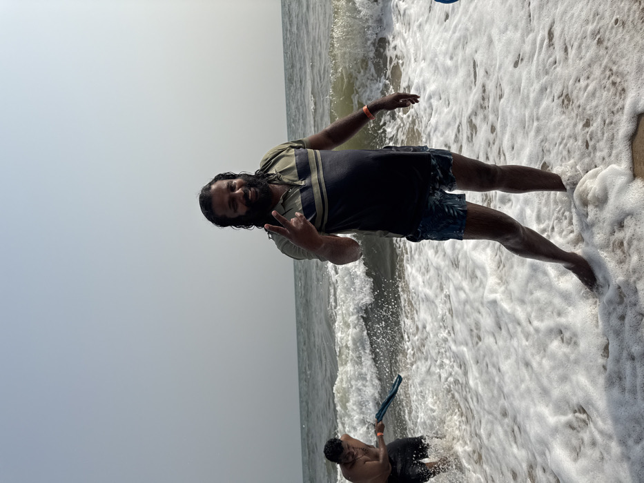 The image displays two adult males at a beach in Mopo Onijebu, Nigeria, with ocean waves and white foam on the shore under a hazy sky.
The central figure, an adult male with long dark hair and a full beard, stands in foamy water approximately mid-calf deep. He wears a striped polo shirt (olive green, dark blue, light green) and dark, patterned swim shorts, with an orange wristband on his right wrist. He smiles towards the viewer, making a peace sign with his left hand.
To the left, a second adult male, bare-chested and wearing dark swim shorts, is partially submerged in deeper water amidst a breaking wave. He holds a dark blue fabric or towel with both hands, appearing to swing or interact with the water, with a discernible expression on his face. The background shows open ocean under a pale, featureless sky.