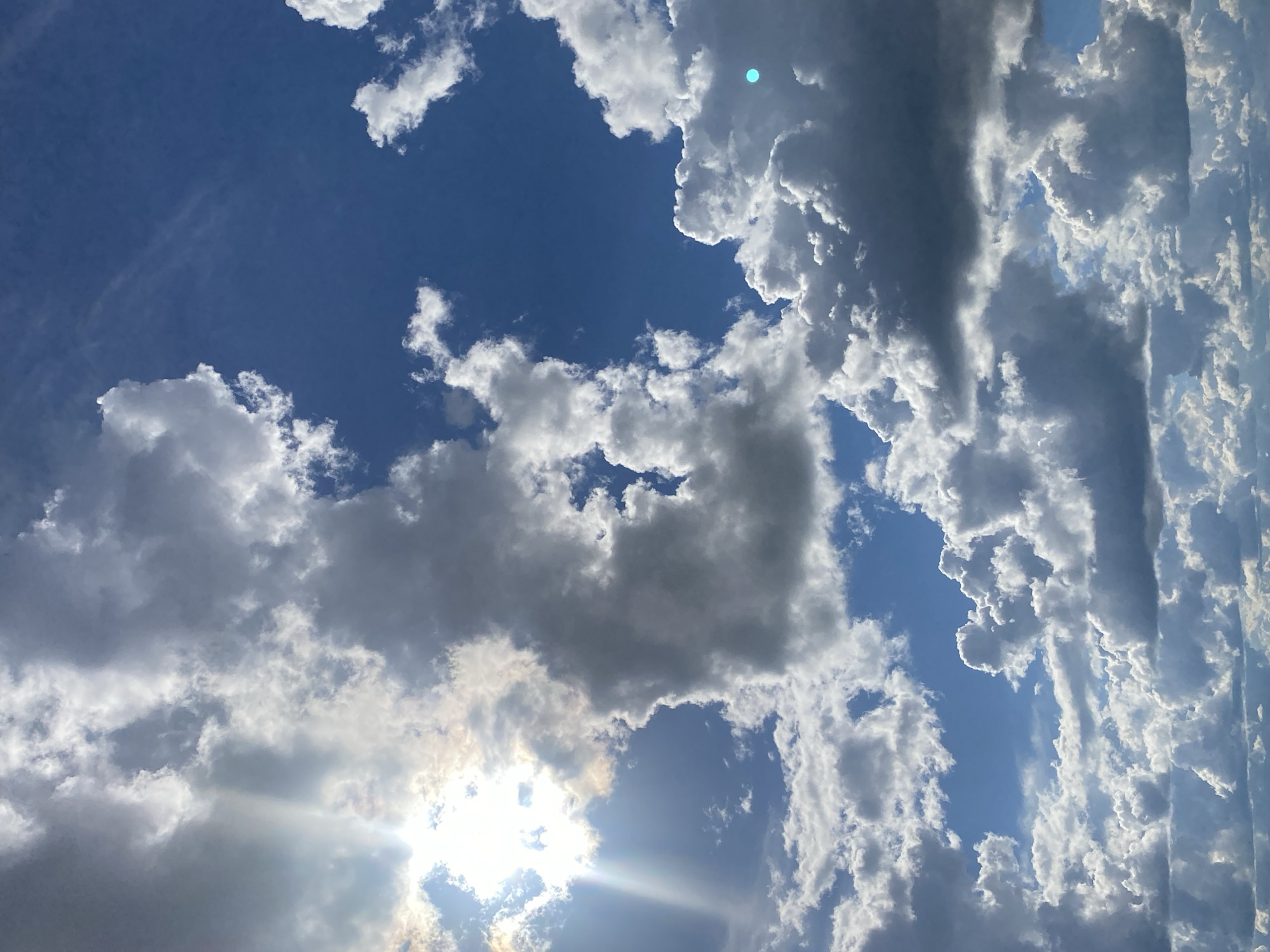 The image shows a daytime sky with a mix of white and gray clouds against a blue backdrop. The sun is visible, partially obscured by clouds, creating a bright glare and sunbeams. The photograph is taken from a low angle, looking upwards. It is not possible to determine the location of the image as Izhevsk, Russia from the image alone.