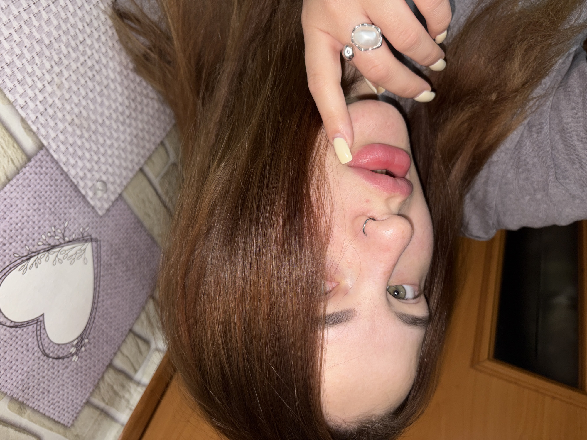A person lies on a surface, with their head at the bottom of the frame. The person has reddish-brown hair, a fair complexion, and light green eyes. They have a nose ring and are wearing a silver ring on their finger. Their fingernails are painted light yellow. The person's hand is near their mouth. In the background, there is a purple and white patterned surface with a white heart design on it. To the right, there is a gray piece of clothing and a wooden door. The location is an indoor setting in the city of Unknown, Unknown.