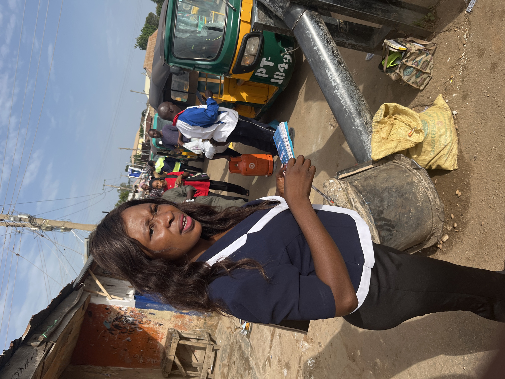 In Gura Top, Nigeria, a woman stands in the foreground, her expression intense as she holds a small stack of brochures. She wears a dark blue jacket with white trim over black pants. Behind her, a busy street scene unfolds. A green and yellow auto-rickshaw, identified by the license plate "PT-1849", is parked amidst other vehicles and pedestrians. An orange gas cylinder sits on the ground near the auto-rickshaw. To the left, a rustic wooden structure with peeling paint and a corrugated metal roof suggests a market stall. The ground is dirt and debris-strewn, illuminated by bright sunlight casting distinct shadows. Power lines crisscross the sky above the urban environment.