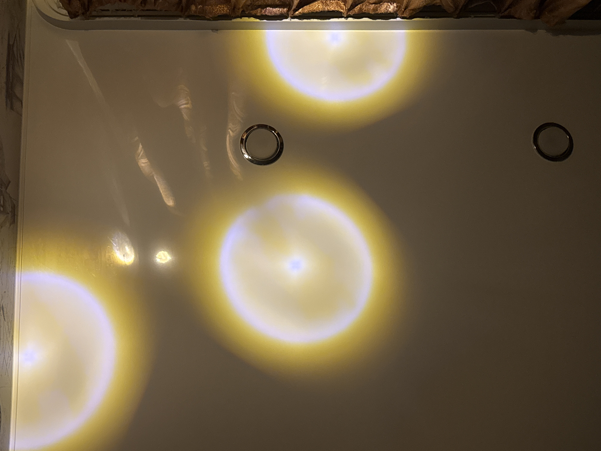 The image captures an indoor scene, showcasing a smooth, highly reflective, white or light-colored surface, likely the base of a bathtub or a shower. Several glowing, circular light reflections with warm, golden-yellow halos and brighter, cooler-toned centers dominate the view, indicative of artificial illumination, possibly from a projector.
Two circular, chrome-like objects are embedded flush with the surface; one is larger and positioned centrally on the left, while a smaller one is located further right. In the upper-left portion of the frame, subtle, elongated, and wavy reflections are discernible, vaguely resembling distorted shapes such as hands or arms, or perhaps patterns from water movement on the reflective surface. At the very top edge, a band of textured, crumpled brown fabric or material is visible, which could be a curtain or valance.
No specific activity is taking place. The artificial nature of the lighting prevents the determination of the time of day. There are no visible weather details or discernible text within the image. While the location context of Chaikovsky, Russia is provided, the image itself offers no specific visual cues to confirm this.
