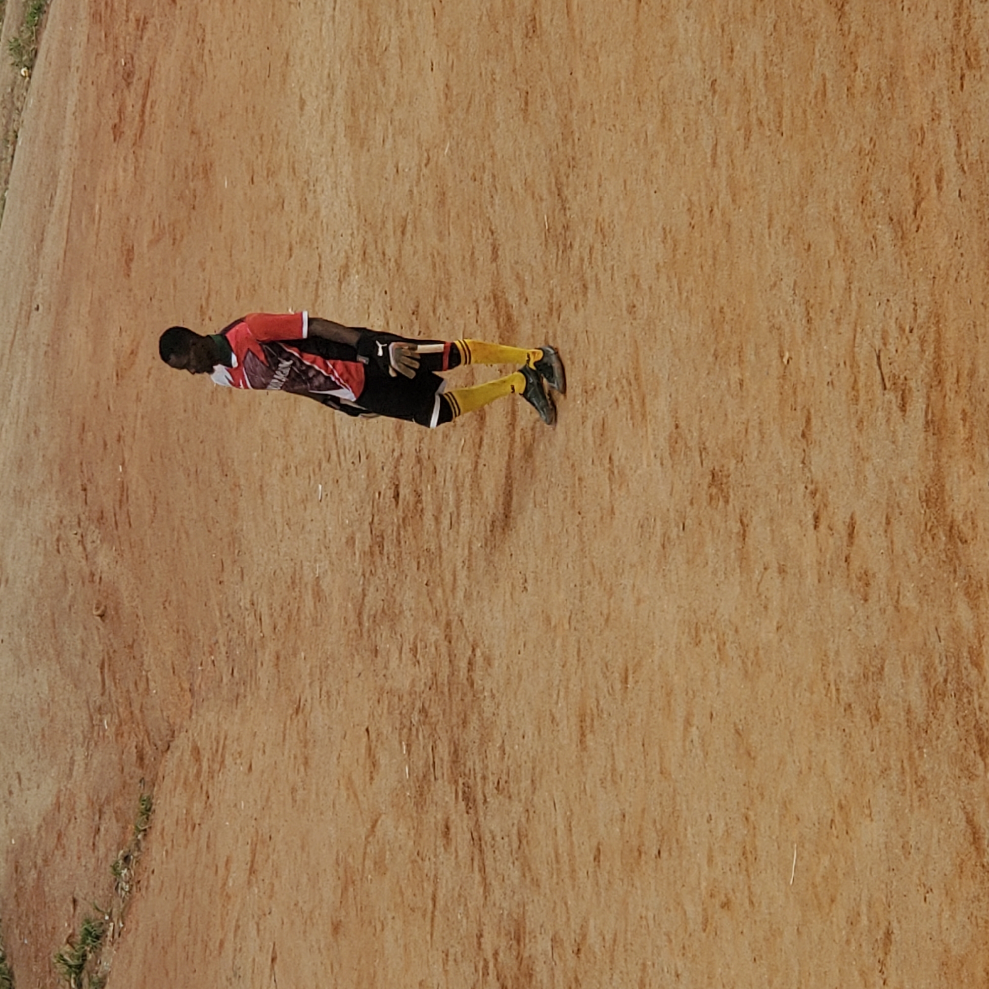 A lone Black male is standing on a dirt field. He is wearing a red and white jersey, black shorts, yellow socks, and dark cleats. He is also wearing a goalie glove on his left hand. The field appears to be dry and dusty, with some patches of grass in the background. The overall impression is that of a casual soccer game being played in a rural or undeveloped area.