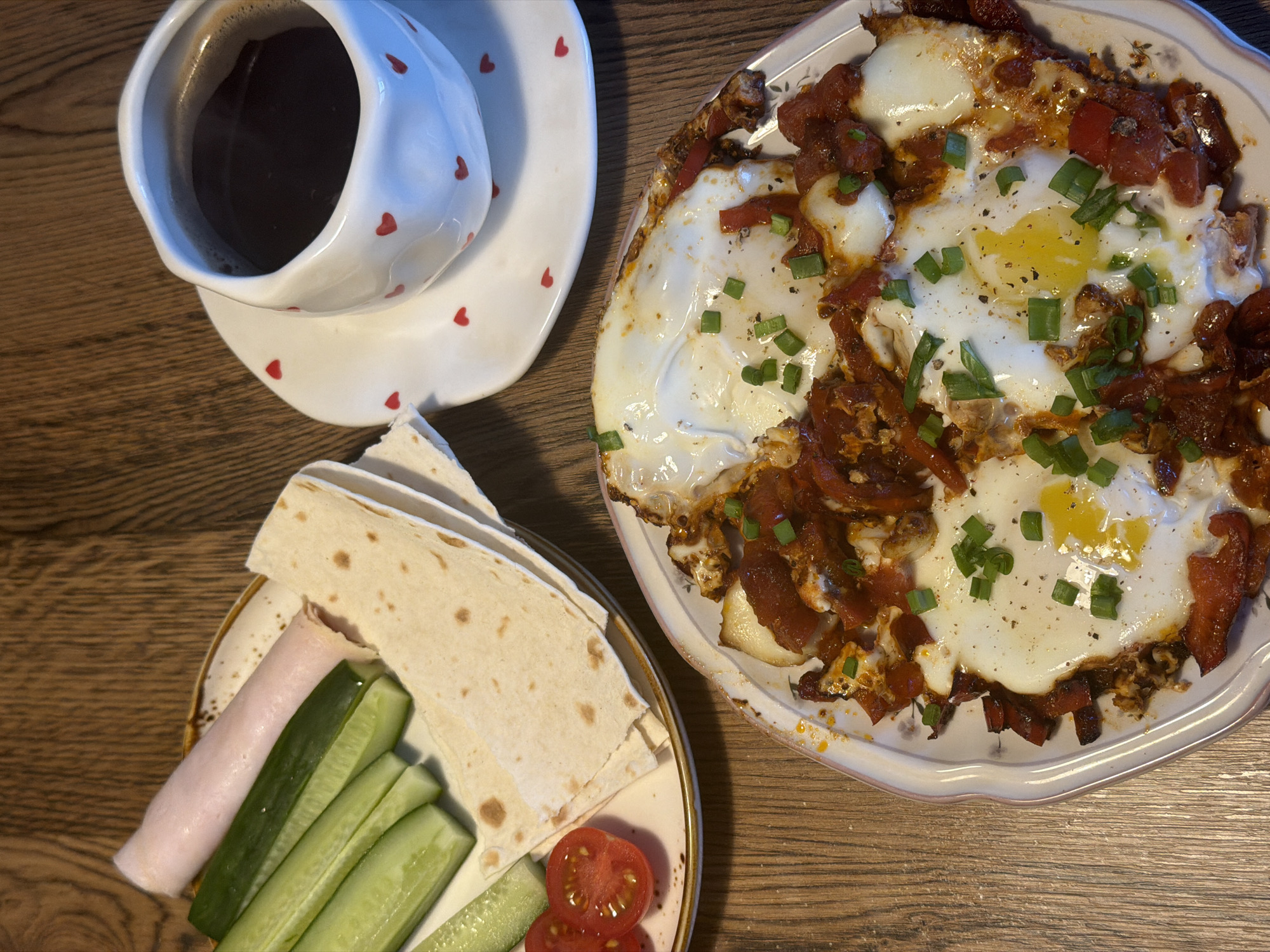 A top-down shot on a wooden surface shows a cup of coffee, a plate with bread, vegetables, and ham, and another plate with shakshuka. The coffee cup is white with red hearts and sits on a matching saucer. The first plate contains folded flatbread, slices of cucumber and tomato, and a rolled slice of ham. The shakshuka plate contains fried eggs in a tomato-based sauce, garnished with chopped green onions and pepper. The image was most likely taken indoors. Location of Perm, Russia is assumed from metadata not present in the image.