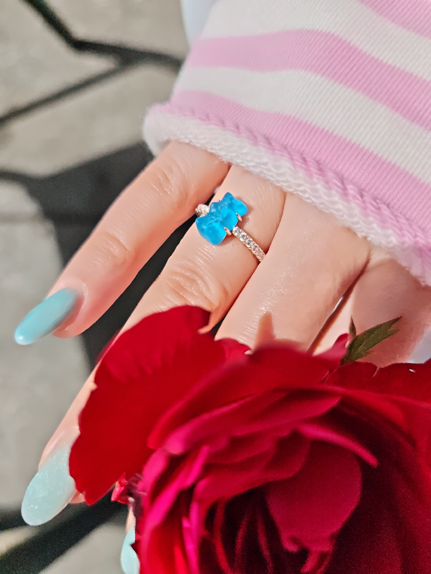A close-up shot focuses on a person's hand adorned with a ring and blue nail polish, partially obscured by a vibrant red rose. The ring features a blue teddy bear-shaped charm surrounded by small, clear stones. The person is wearing a pink and white striped long-sleeved shirt with a white fuzzy trim. The background appears to be an outdoor setting with a blurred, greyish surface that suggests paving stones. The lighting indicates it is daytime. There are no discernible elements that would indicate a specific location within the city of Schwerin, Germany.