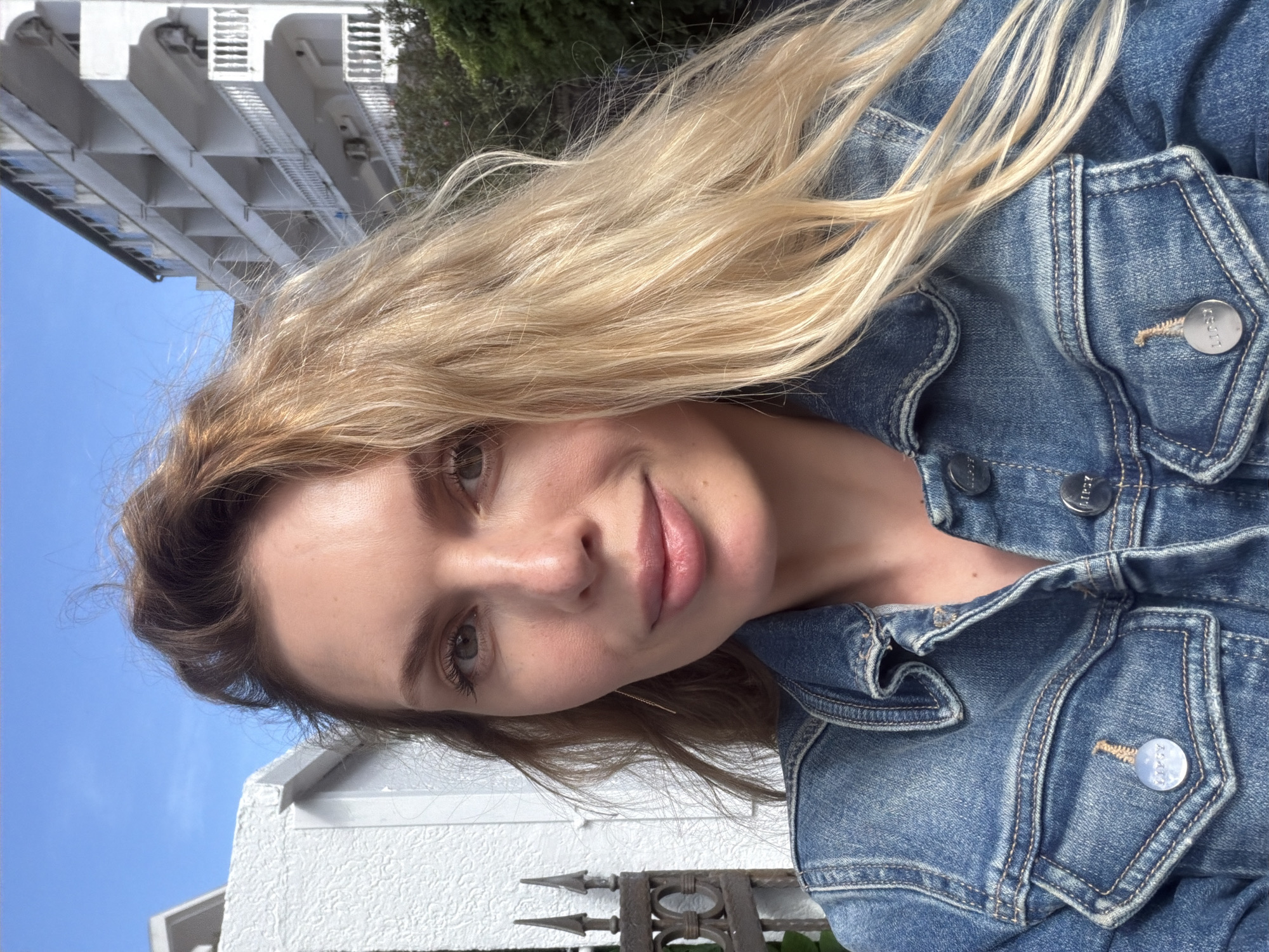 A woman with long, blonde hair is wearing a denim jacket in an outdoor setting. A white building with multiple levels and balconies is partially visible in the upper left background. The sky is blue. A white structure and wrought iron fence appear in the lower background. The exact location in Budva, Montenegro cannot be determined with only this image.