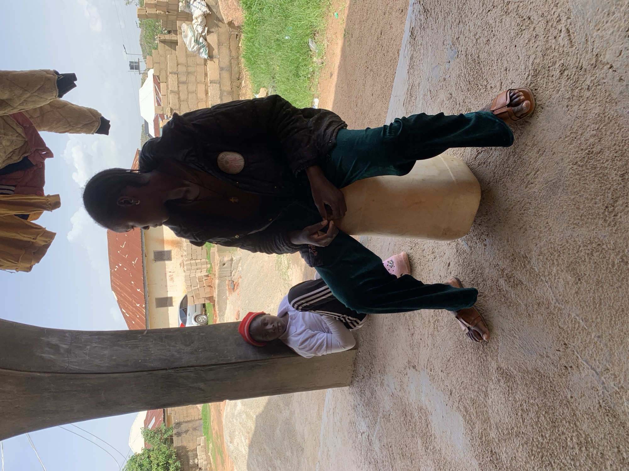 Two people are sitting outdoors in what appears to be a rural or semi-urban setting in Jos, Nigeria. The person in the foreground, leaning against a large, light-colored cylindrical object, is wearing a dark jacket, dark green trousers, and brown sandals. They are looking down, with their hands clasped in their lap. The second person is lying down, resting their head on a concrete ledge, and is wearing a white hooded top with black stripes on the sleeves, dark trousers, and a red hat. They appear to be asleep or resting.
The scene is outdoors with a clear sky, suggesting daytime. In the background, there are brick buildings, some with corrugated metal roofs, and some construction materials. A silver car is visible parked near one of the buildings. Green grass can be seen in some areas. The ground is made of concrete and dirt. The overall impression is one of a humble living environment. There is no visible text in the image. The mood seems relaxed, possibly indicating a break or a moment of repose.