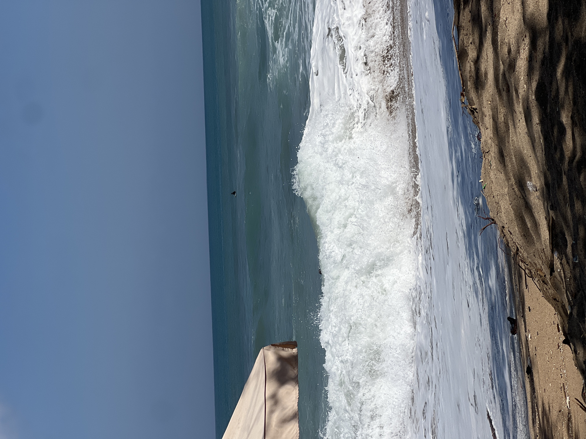 An eye-level, medium shot captures a bright, sunny day at Kuta Beach, Bali, Indonesia. The scene is dominated by the ocean, with a wave in the foreground breaking into white foam as it crashes onto the shore. In the distance, the turquoise water stretches to the horizon, meeting a clear, pale blue sky. A single, dark figure, possibly a person or a large bird, is visible on the water in the far distance. To the left of the frame, the corner of a beige beach umbrella or cabana is visible, casting a shadow on the sand. The foreground consists of wet sand with debris and shadows of palm fronds cast from an unseen source. The overall impression is one of a tranquil beach setting with the dynamic energy of the ocean.