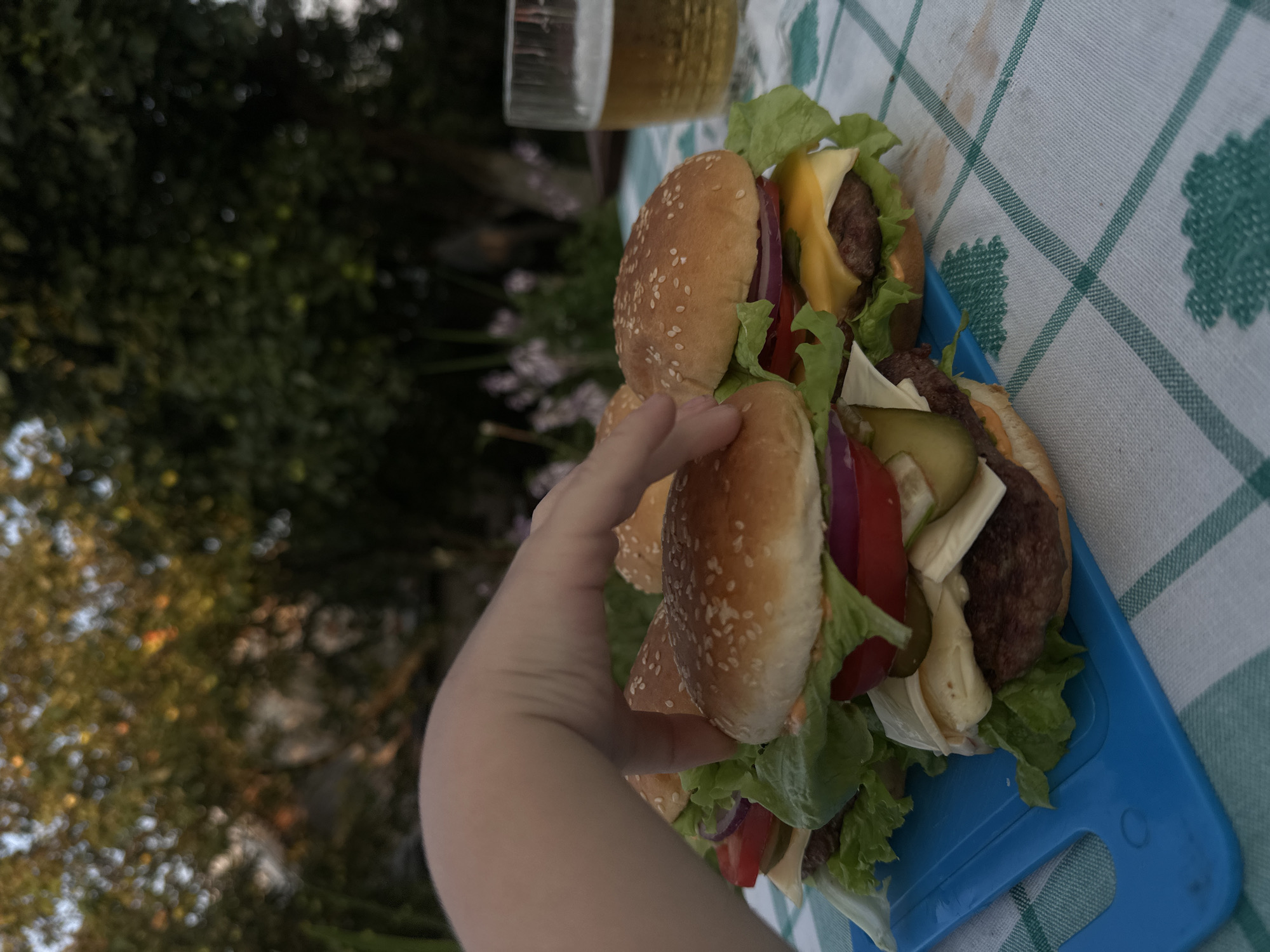 An overhead close-up shot shows a person's hand reaching for a hamburger in an outdoor setting, likely in Galevo, Russia. There are several hamburgers on a blue tray, with a glass of beer in the background. The hamburgers are elaborately dressed with lettuce, tomatoes, onions, pickles, and cheese. The background is out of focus, showing trees and foliage, suggesting a park or backyard setting. The table is covered with a green and white checkered tablecloth.