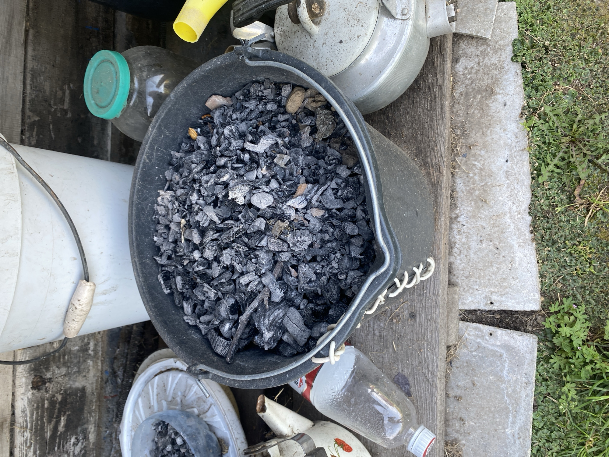 The image shows an outdoor scene likely in Krupki, Belarus. In the foreground, a large black plastic bucket is filled with charcoal. To the left of the bucket is a glass bottle with a partially removed red and white label, and a small white kettle with a strawberry design. Behind the bucket, a large white bucket is visible, and to its right, a glass jar with a green lid. Further to the right, a yellow funnel is partially visible, and a metal teapot with a gray lid. The objects are arranged on a wooden surface, with concrete slabs and grass visible below. The background consists of a wooden wall.