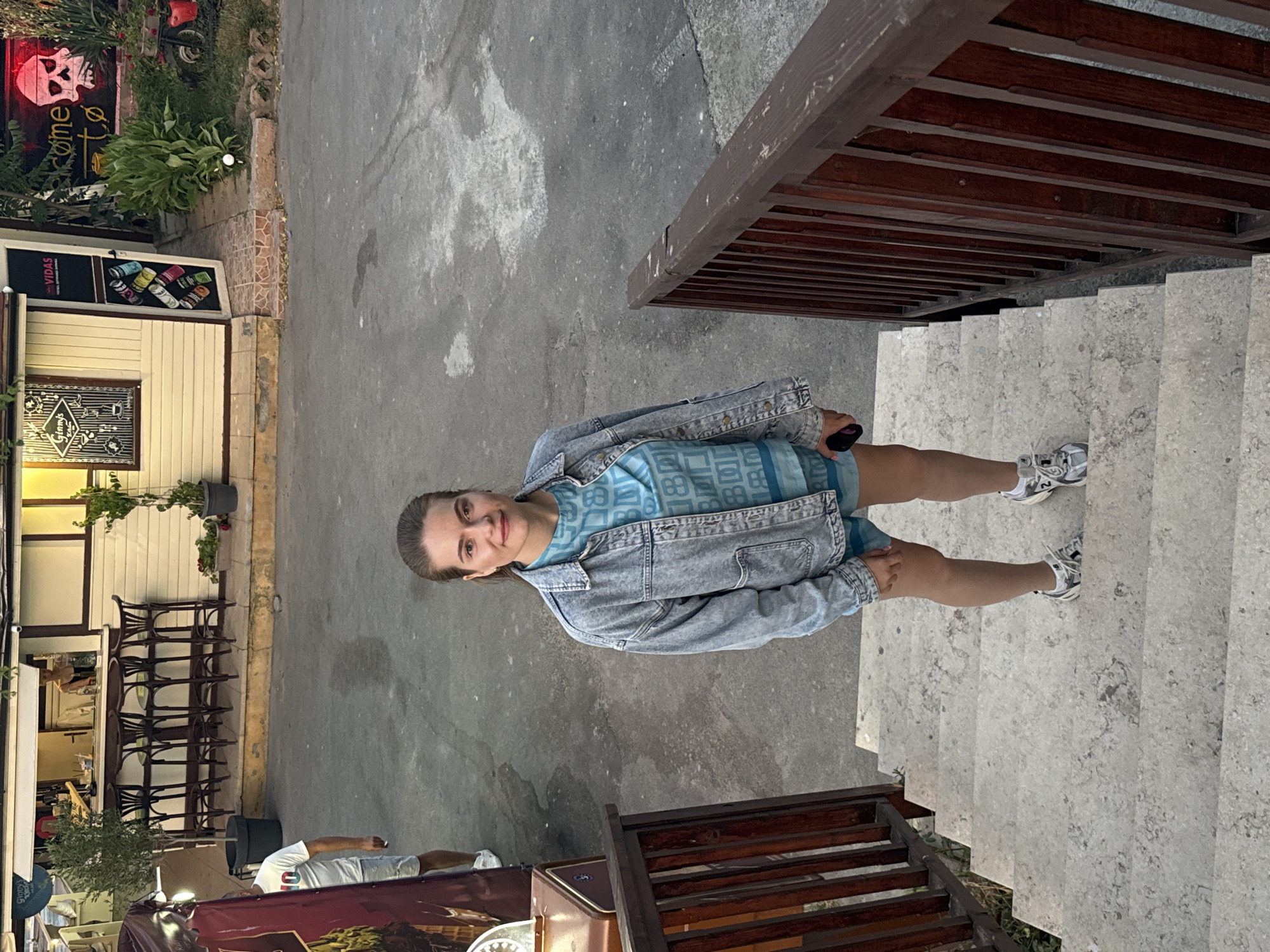 A young woman is standing on outdoor concrete steps. She is wearing a light blue denim jacket over a blue patterned t-shirt and blue shorts. She has on white sneakers. The woman is looking towards the camera and smiling.
In the background to the left, there is a building with a white facade and a dark sign that says "VIDAS". Next to it is a black sign with a red skull and the word "come". There are some chairs and tables outside the building. The setting appears to be an outdoor cafe or bar. The ground is paved with asphalt. To the right of the woman, there is a wooden railing and more concrete steps.
The lighting suggests it might be daytime, possibly late afternoon given the soft light. The overall atmosphere seems relaxed and casual. The location is identified as Sozopol, Bulgaria. There are no clear indicators of weather, but the scene appears dry.
