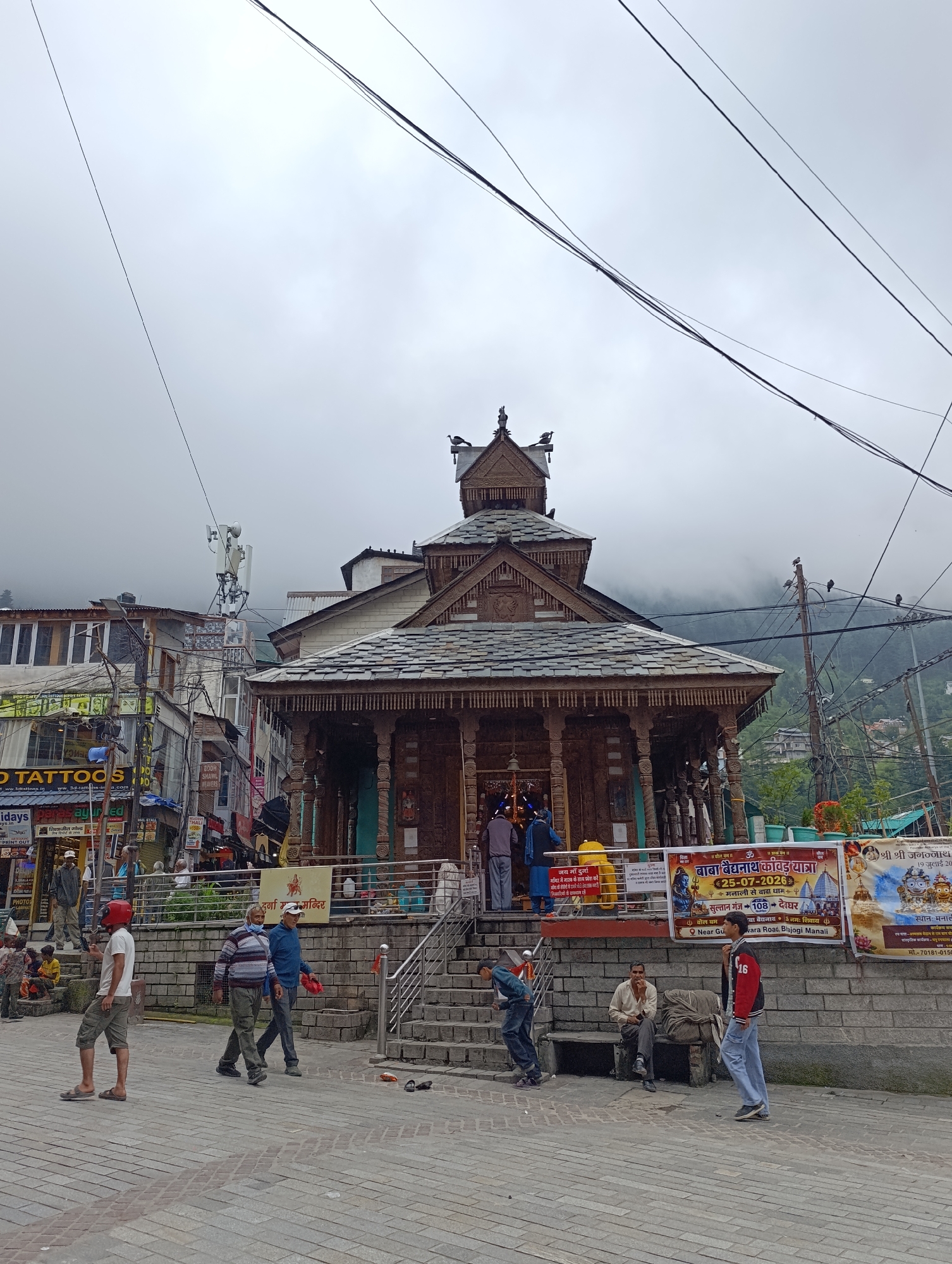 The image depicts a street scene in Manali, India, with a prominent wooden temple in the center, characterized by intricate carvings and a slate roof. The temple has a raised entrance with stone steps. In front of the temple, there is a paved walkway with several individuals. To the left of the temple, a row of buildings with shops is visible, including signs for "3D TATTOOS" and other businesses. Electrical wires and poles are prevalent throughout the scene, indicating urban infrastructure. In the background, misty mountains can be seen, characteristic of the Manali region. Several people are walking, standing, or sitting on benches. One person in the foreground is wearing a red helmet. Banners with text in Hindi are displayed near the temple, advertising events or religious services.