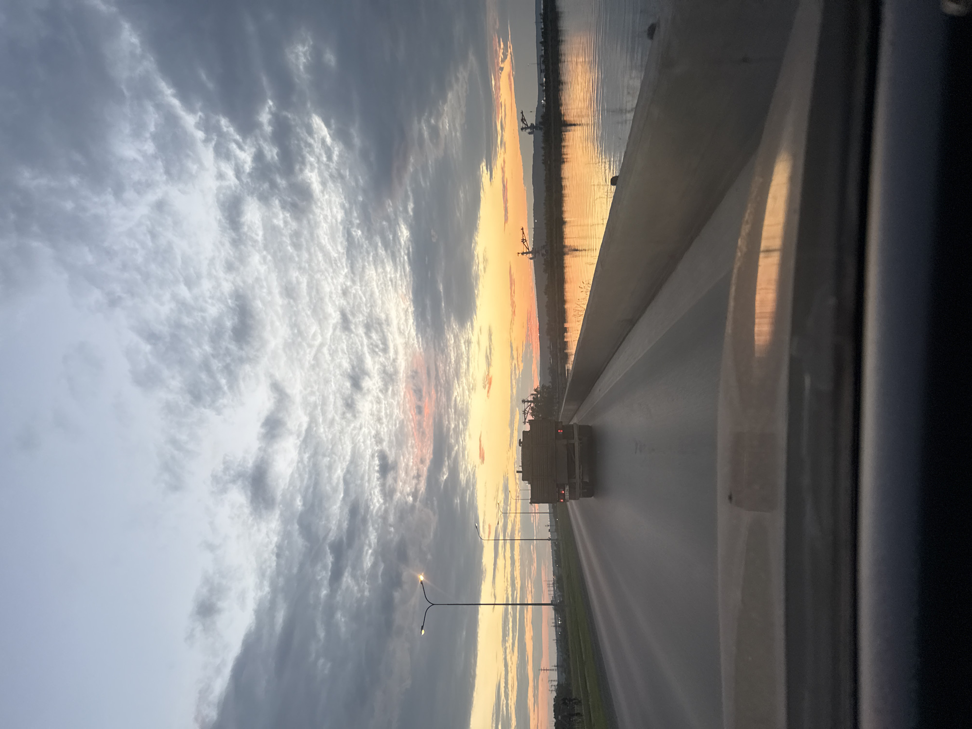 The image captures a view from a vehicle traveling on a road that appears to be a bridge or elevated causeway, likely near the city of Chaikovsky, Russia, given the context of the request. The time of day is sunset, indicated by the warm orange and pink hues in the sky and the reflections on the water to the right of the road.
On the road, a tan truck is driving away from the camera. Further ahead, two streetlights illuminate the roadway. To the left of the road, a grassy median separates it from what appears to be fields or undeveloped land, with some power lines visible in the distance.
The body of water to the right is calm, with a clear reflection of the sunset and some industrial-looking structures or cranes on the distant shoreline. The sky is filled with a mix of dark gray and lighter, textured clouds, with the brightest light concentrated near the horizon. The foreground of the image shows the interior of the vehicle, specifically the dashboard and windshield, with reflections of the sky and road visible.