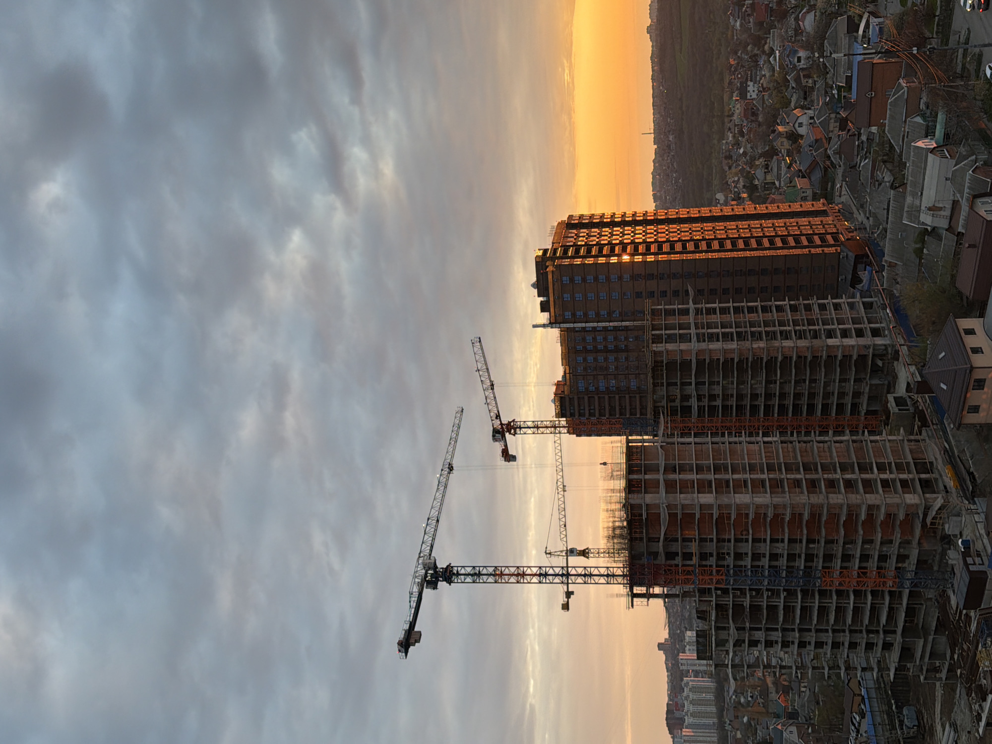 The image shows a construction site in a city, with several high-rise buildings under construction and cranes in operation. The sun is setting, casting an orange glow on the buildings and sky. In the background, there are residential buildings and a wooded area. The sky is filled with clouds, suggesting a cloudy or overcast day.