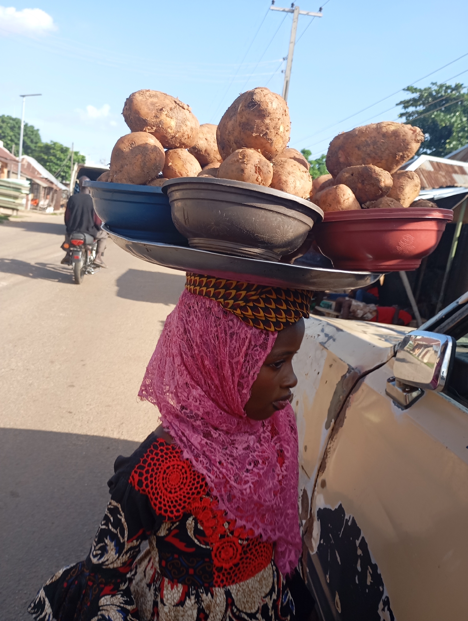 In Shendam, Nigeria, a young person is pictured carrying a load of root vegetables, likely yams or sweet potatoes, balanced on their head. The vegetables are contained within three stacked plastic bowls, with a metal tray placed between the lower two. The individual is wearing a pink lace headscarf and a colorful patterned dress. They are standing next to a beige vehicle, with a side-view mirror visible. In the background, a street scene unfolds with buildings, trees, a power pole, and a person riding a motorcycle. The lighting suggests it is daytime.