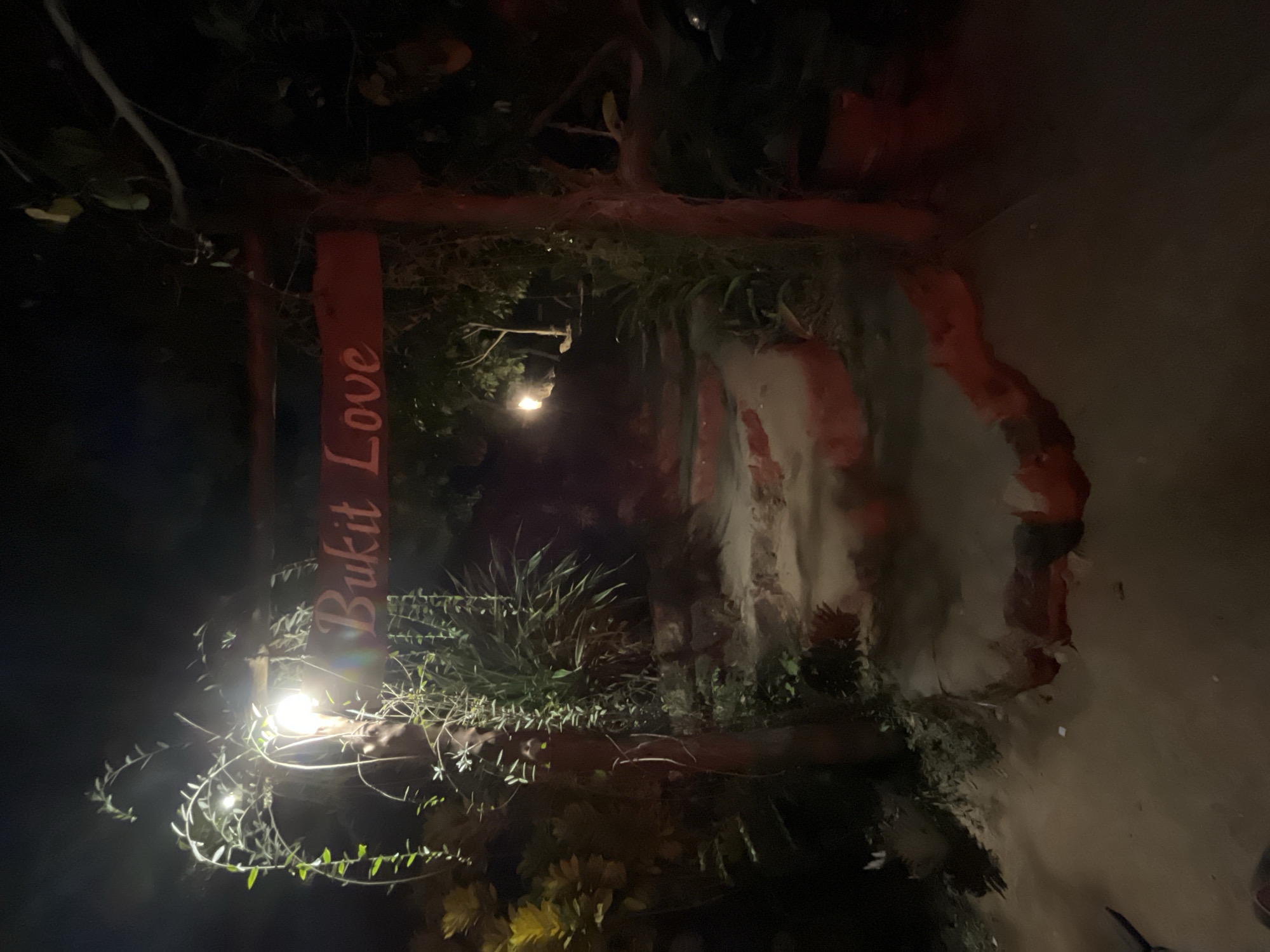 The image depicts a nighttime scene of a pathway in Karimunjawa, Indonesia. A wooden sign with red lettering reading "Bukit Love" is illuminated by a light fixture hanging from an overhead branch, casting a warm glow on the surrounding foliage. The pathway itself is constructed of stone steps, with red-painted edges, leading upwards into the darkness. Lush green plants and trees frame the path, with some hanging vines adding to the natural ambiance. The overall impression is one of a tranquil and inviting entrance to a scenic location.