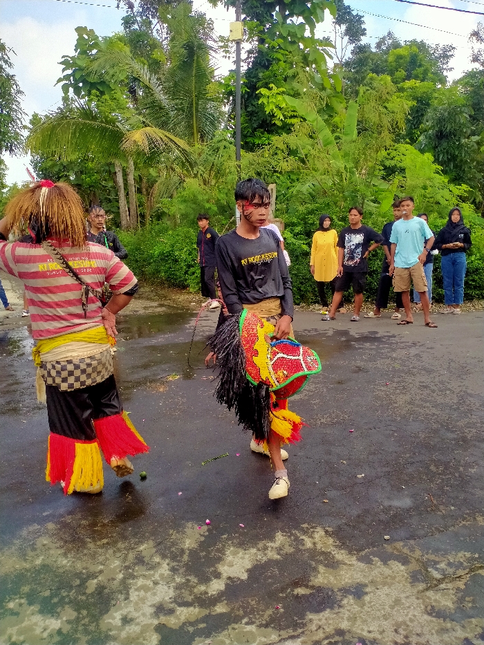 The image shows a group of people standing on a paved road in Kecamatan Kedawung, Indonesia (latitude: -7.514, longitude: 111.03). There are two people in the foreground. The person on the left is wearing a red and white striped shirt with a yellow and black skirt. They have a black strap around their waist. The person on the right is wearing a black shirt and black pants. They have a brightly coloured mask made of red, yellow, and green. There are other people in the background, wearing various attire. The image is taken from a slightly elevated angle. The road is wet, and there are pink flower petals scattered on it. The background includes lush green trees and a light pole.