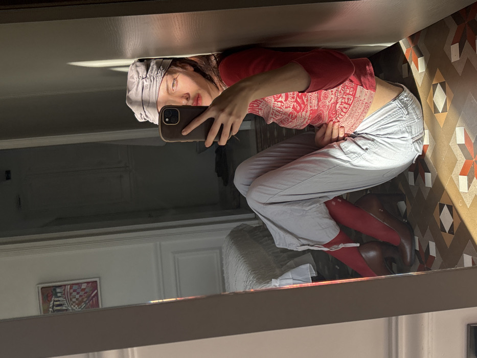 A female individual is seated on a geometric patterned tile floor, consistent with traditional hydraulic tiles found in residences in Barcelona, Spain. She leans against a light-colored wall or doorframe on the right side of the frame, capturing her reflection in a full-length mirror positioned to her left. Her face is partially obscured by a dark-colored smartphone, held in her right hand as she takes a mirror selfie. She wears a light gray ruched cap, a red long-sleeved top with light-colored patterns on the torso, and light blue or gray cargo-style pants. Red socks or tights are visible on her legs, terminating in dark brown high-heeled shoes. Direct sunlight illuminates her face, the floor, and casts a sharp shadow on the wall to her right. The mirror reflection reveals a room with a white wall, decorative paneling, a bed with a white ruffled bedspread, and a framed piece of abstract artwork featuring red, white, blue, and yellow elements. Beyond a partially open door in the background, a dimly lit area suggests a utility space or bathroom, with a faint view of a toilet fixture.